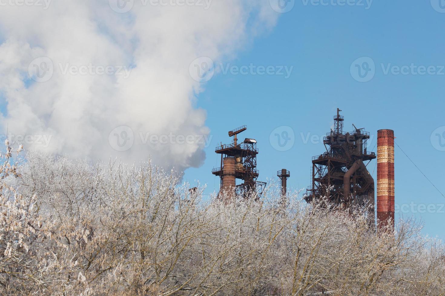 metallurgical plant with cloud of pollution exhaust with frozen winter bush in foreground at daylight photo