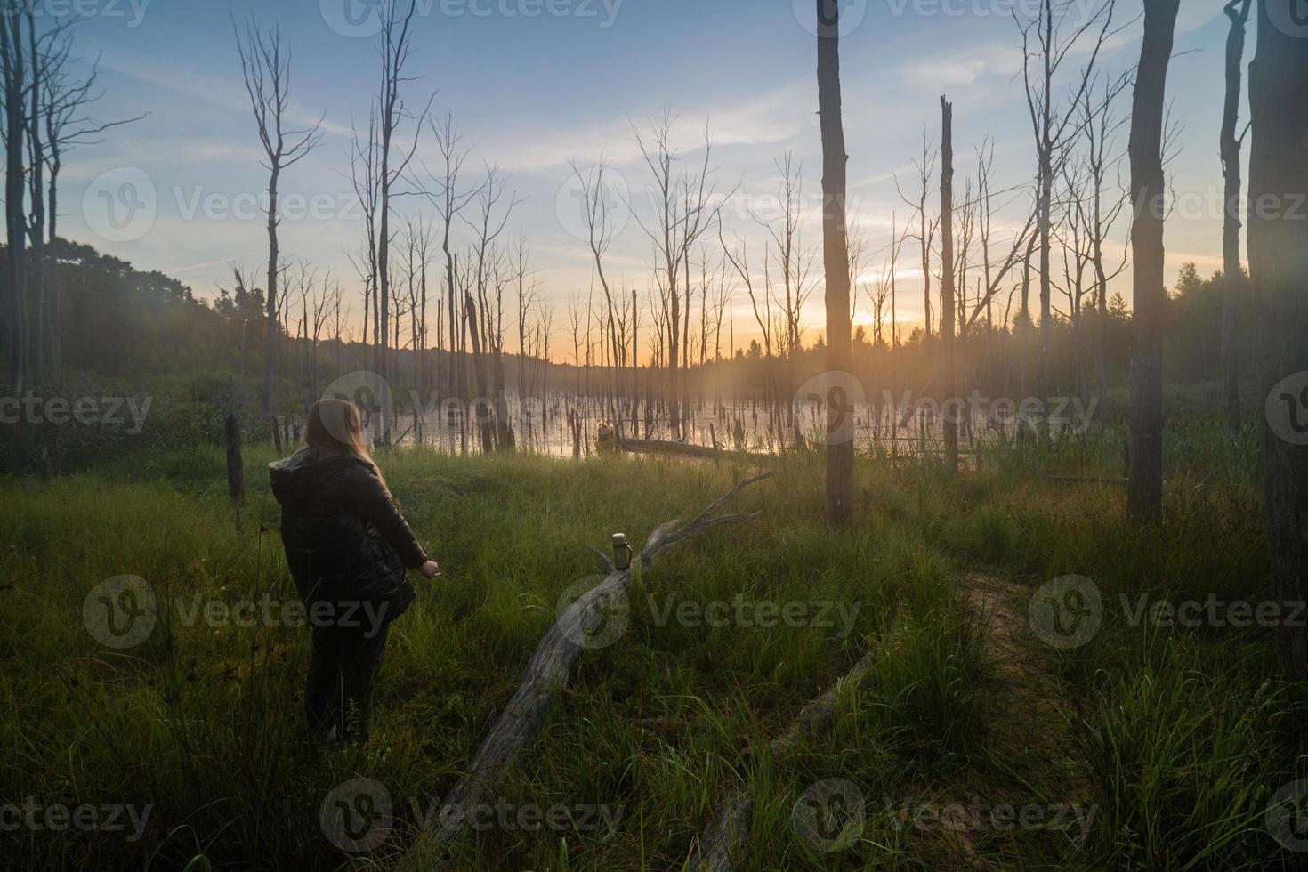 young blonde caucasian woman smoking on the edge of morning summer swamp with dry gray tree trunk - dreamy wide angle shot photo
