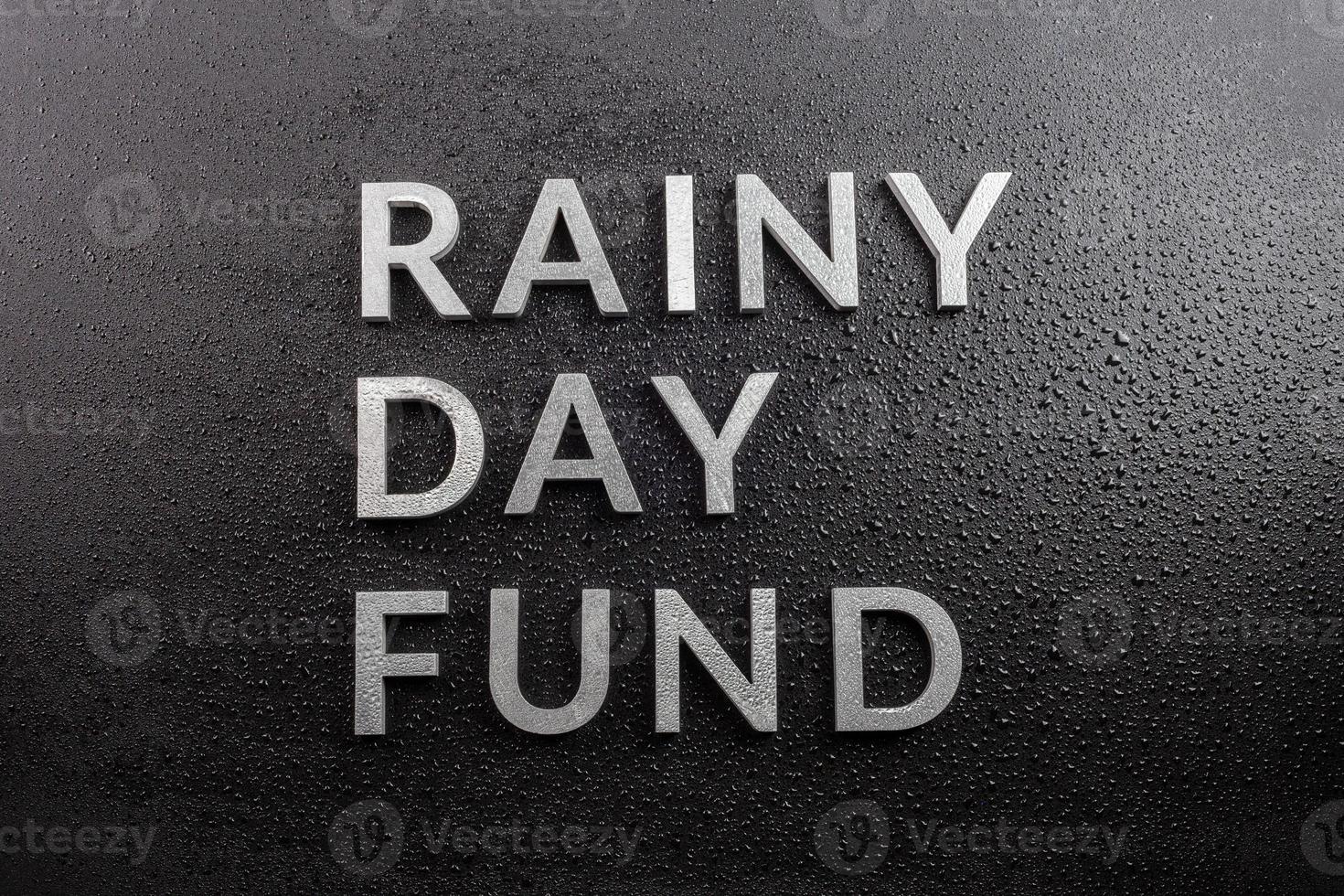 the words rainy day fund laid with silver metal letters on black background, covered with water drops, flat lay perspective, directly above photo