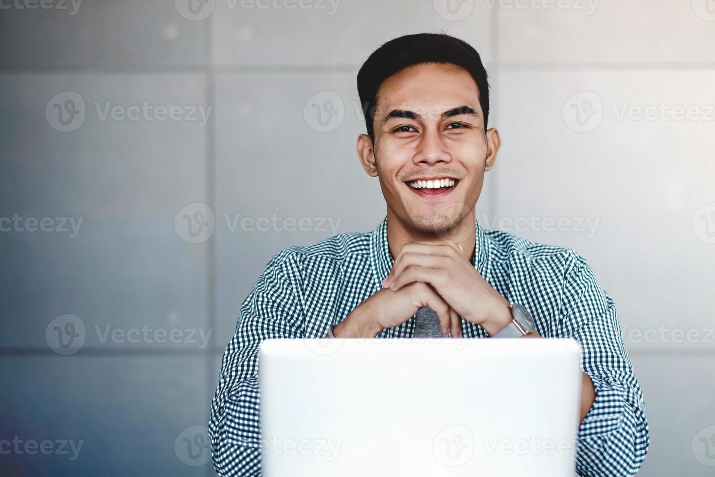 Happy Young Asian Businessman Working on Computer Laptop in his Workplace. Hands on Chin, Smiling and looking at Camera photo