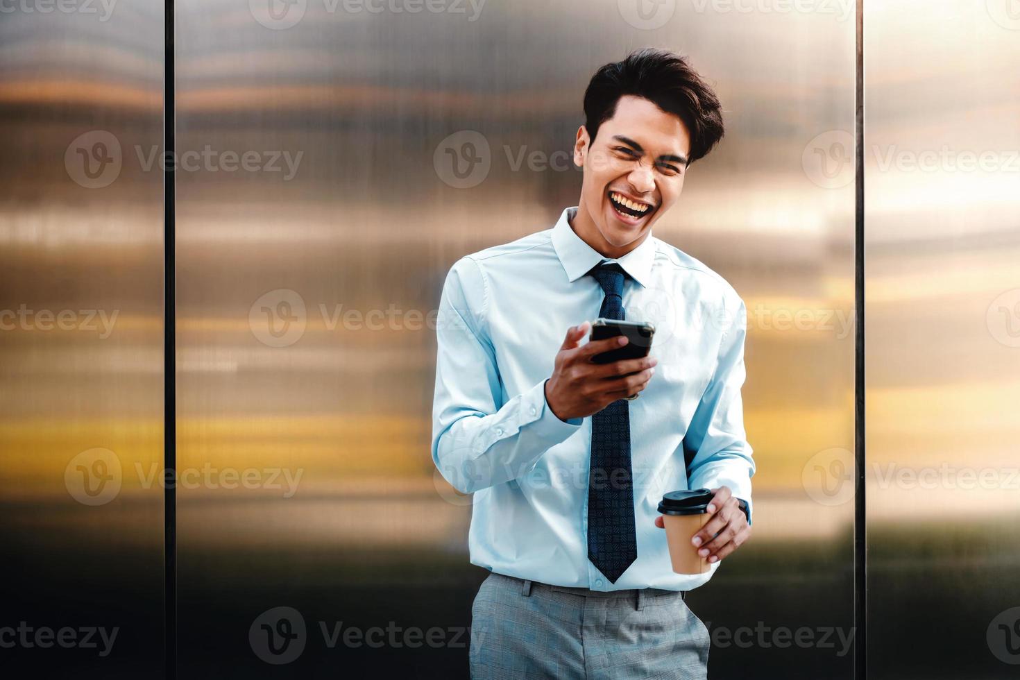 Portrait of a Happy Young Businessman Using Mobile Phone in the Urban City. Lifestyle of Modern People. Front View. looking at Camera. Standing by the Wall with Coffee Cup photo