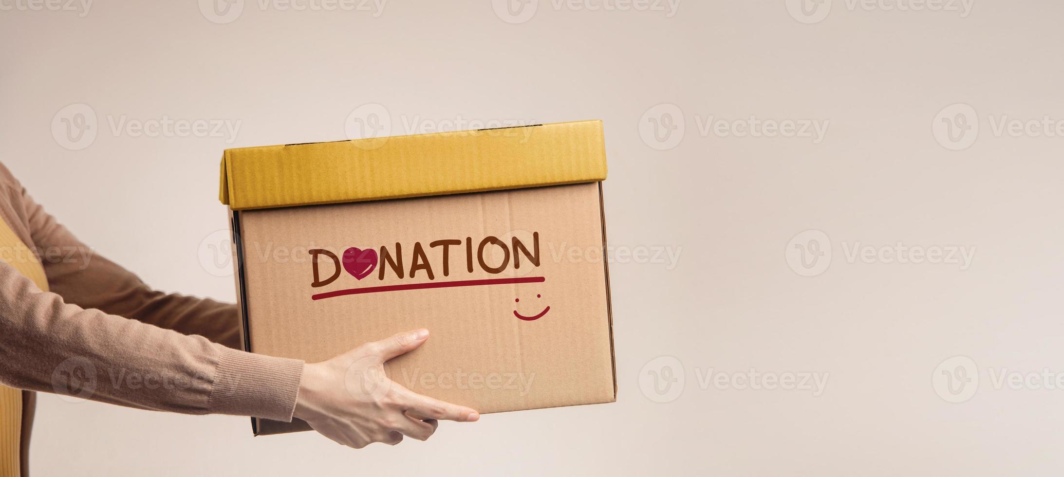 concepto de donación. mujer con caja de cosas para donar con etiqueta de donación, sonriendo y corazón. de pie contra la pared. vista lateral foto