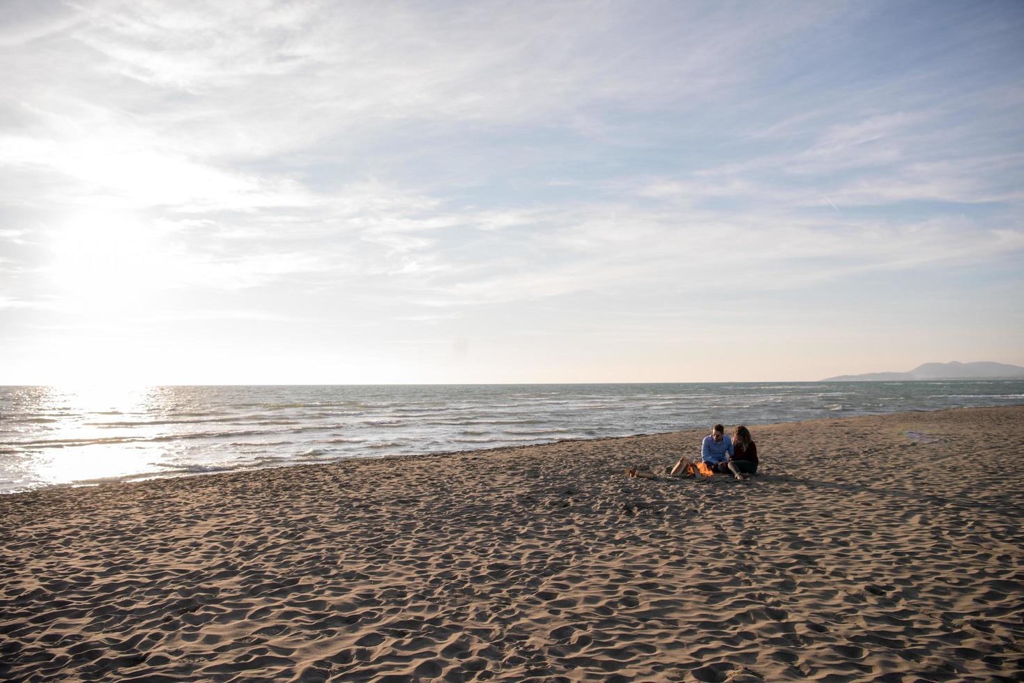 pareja joven sentada en la playa al lado de una fogata bebiendo cerveza foto