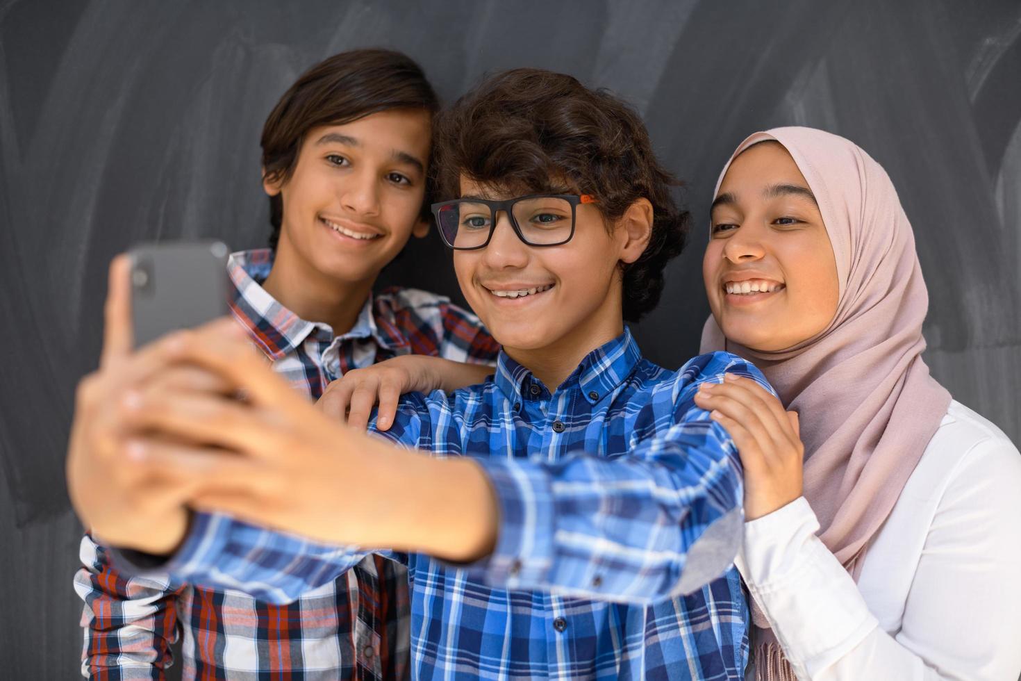 Group of arab teens taking selfie photo on smart phone with black chalkboard in background. Selective focus