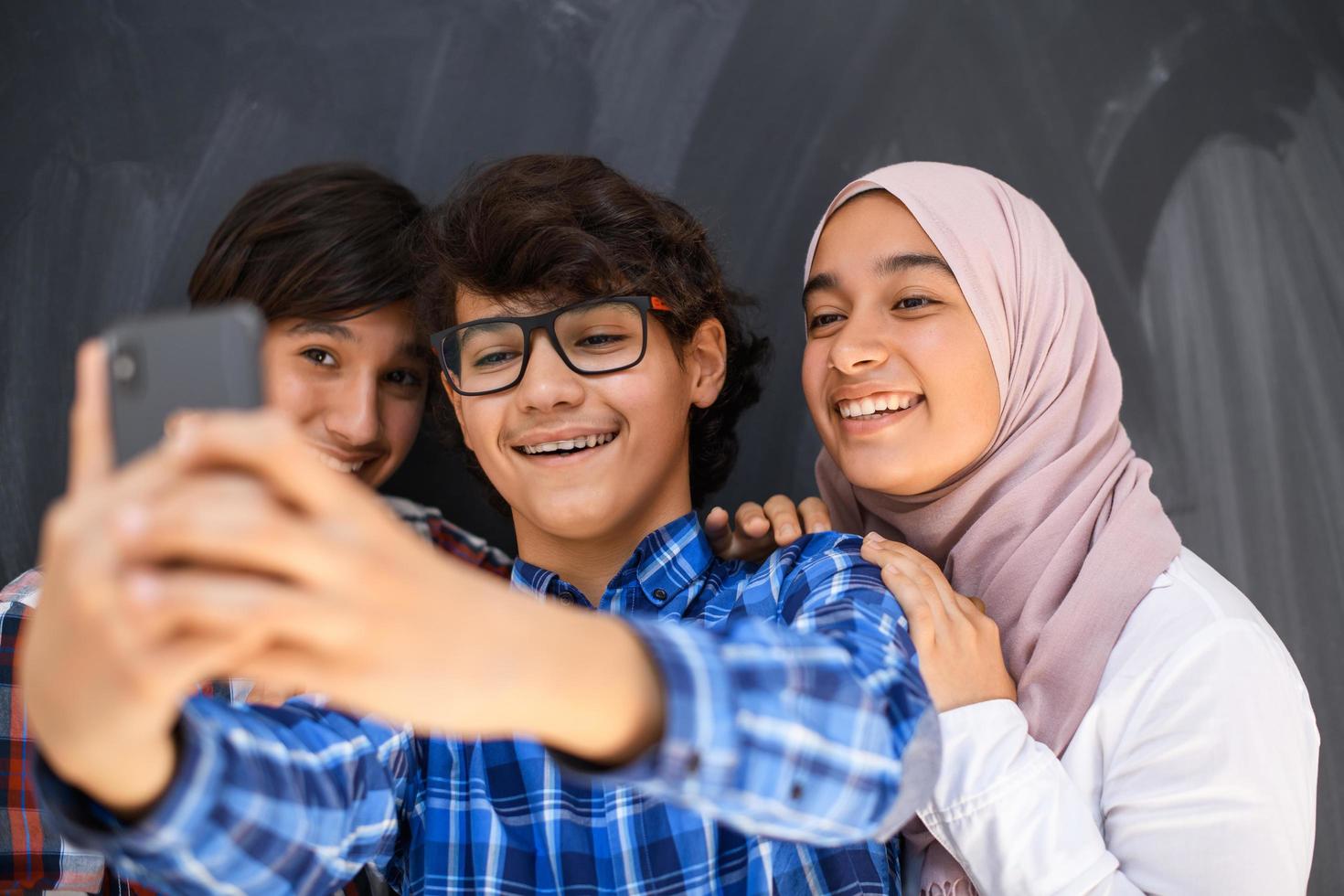 Group of arab teens taking selfie photo on smart phone with black chalkboard in background. Selective focus