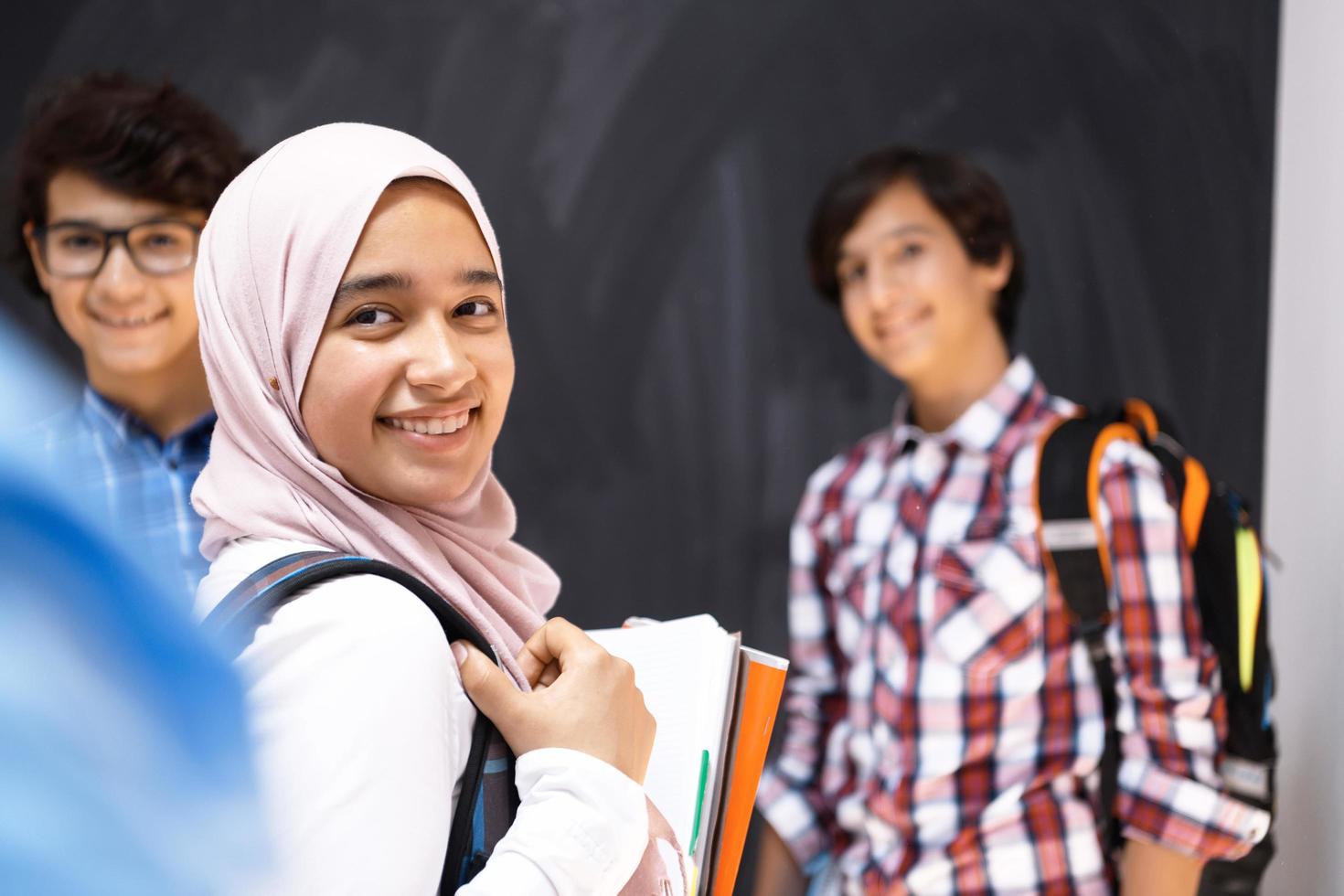 Arabic teenagers, students group portrait against black chalkboard wearing backpack and books in school.Selective focus photo