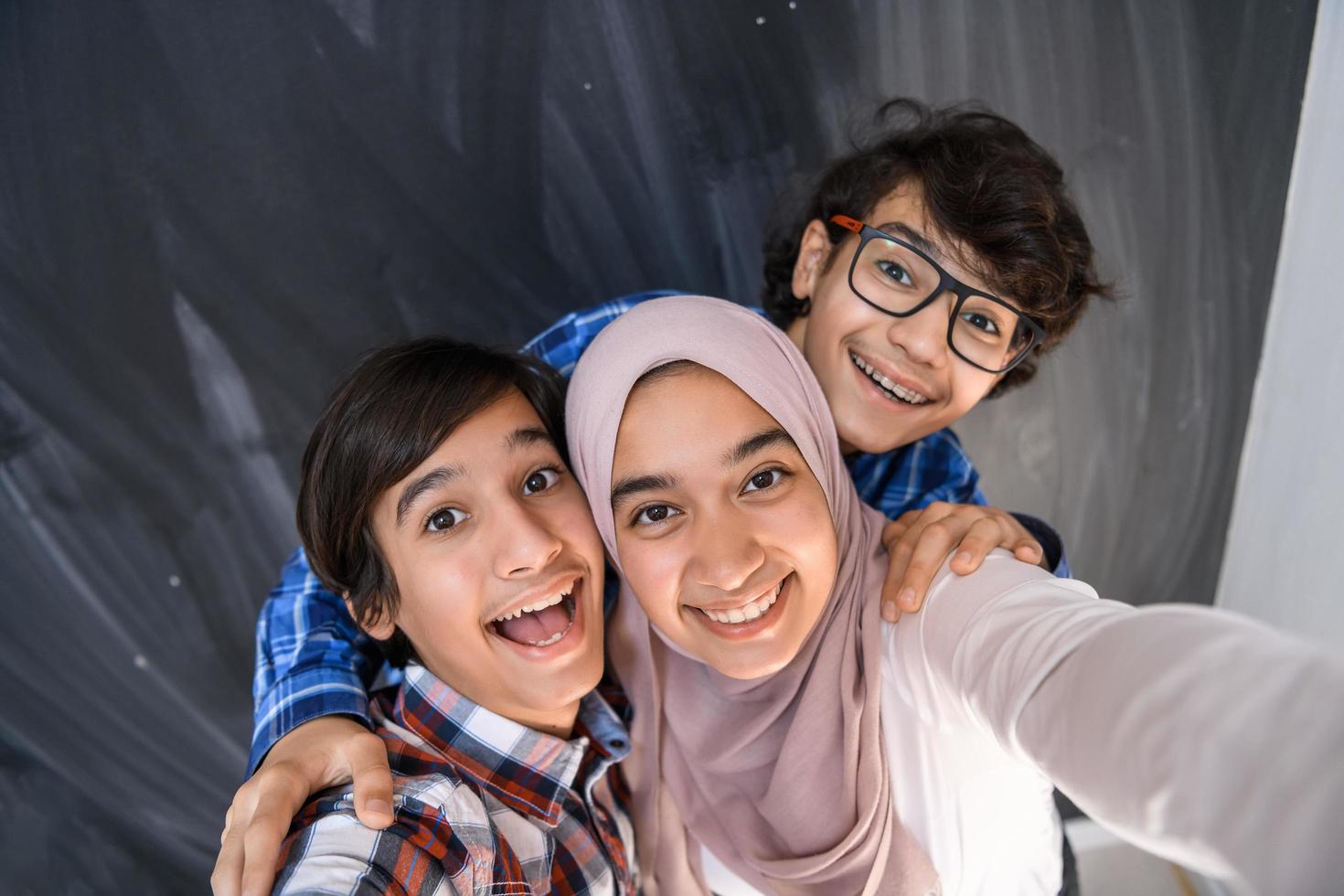 Group of arab teens taking selfie photo on smart phone with black chalkboard in background. Selective focus