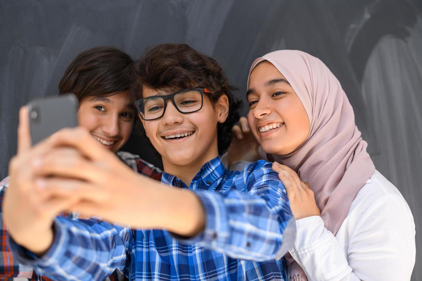Group of arab teens taking selfie photo on smart phone with black chalkboard in background. Selective focus