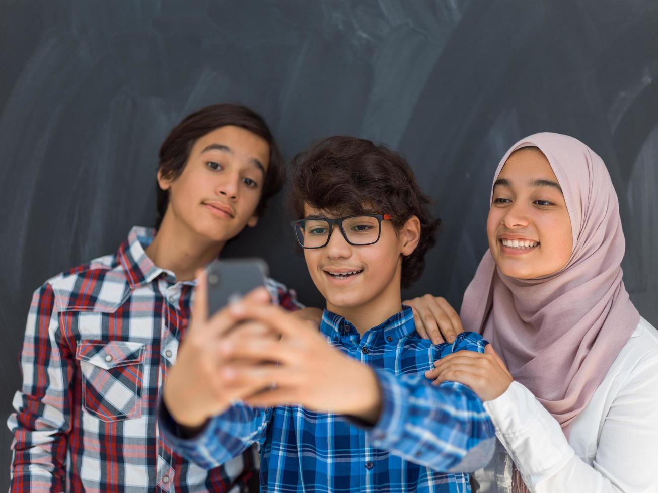 group of arab teens taking selfie photo on smart phone with black chalkboard in background