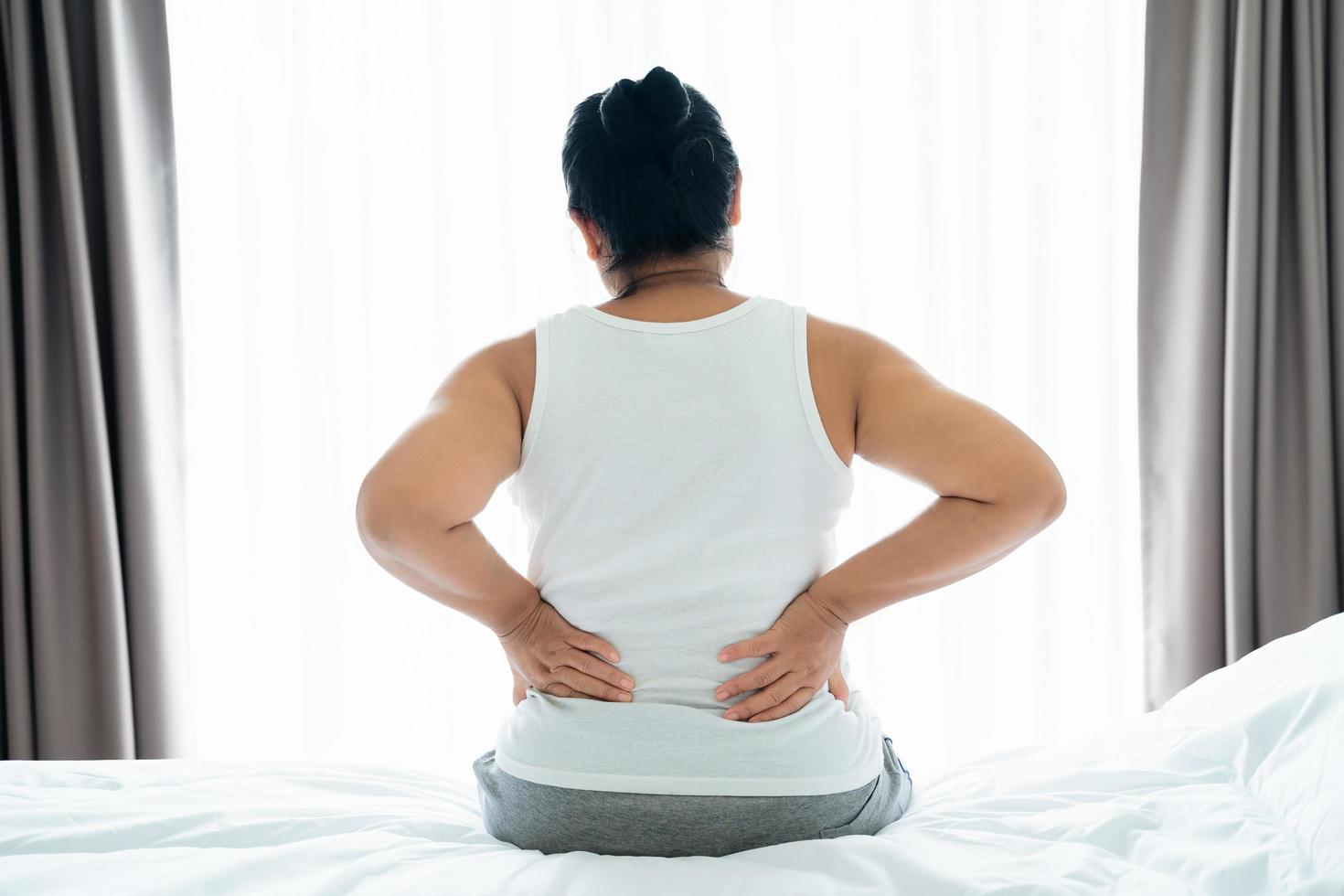 Adult Asian woman is sitting on the bed and holding her lower back suffering from injured back. Health care and back pain concept. photo