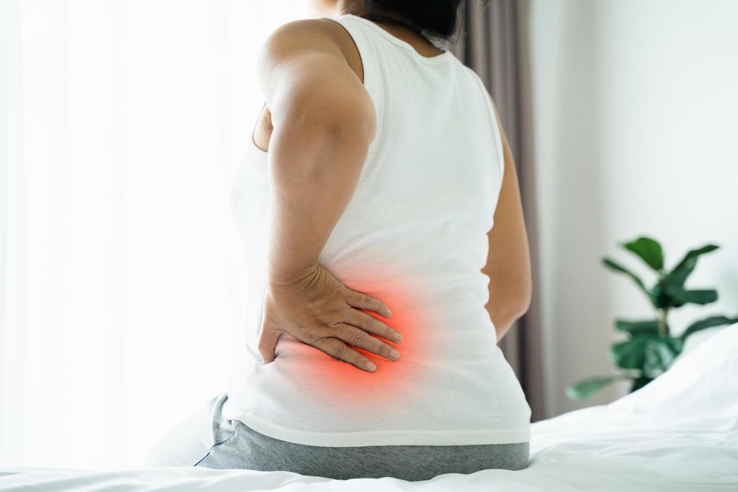 Adult Asian woman is sitting on the bed and holding her lower back suffering from injured back. Health care and back pain concept. photo