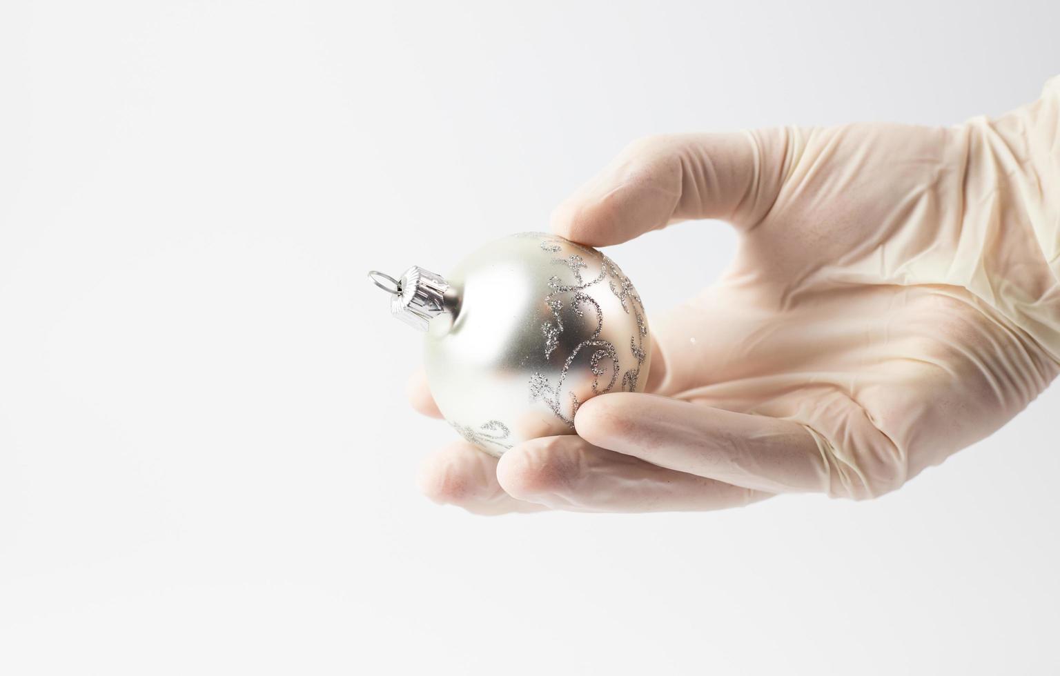 A silver Christmas bauble in a hand wearing a protective glove. Symbolism of the holidays during the pandemic. White background. photo