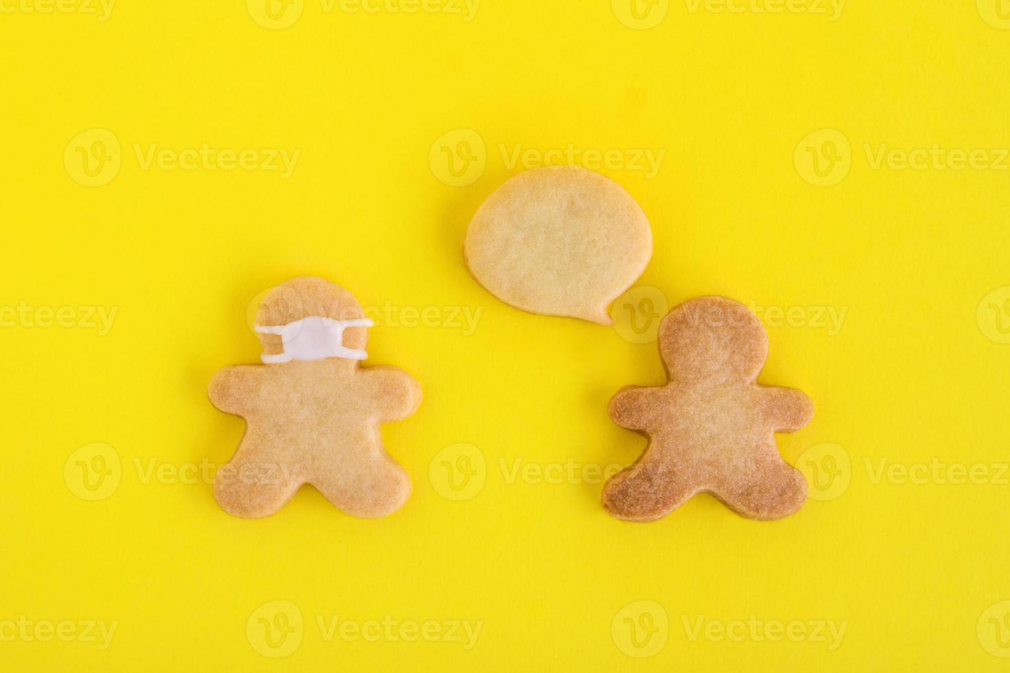 Homemade cookies in shapes of people with callout cloud and with face medical mask on yellow background, top view. Sweet shortbread with white glaze. Social distancing concept. photo