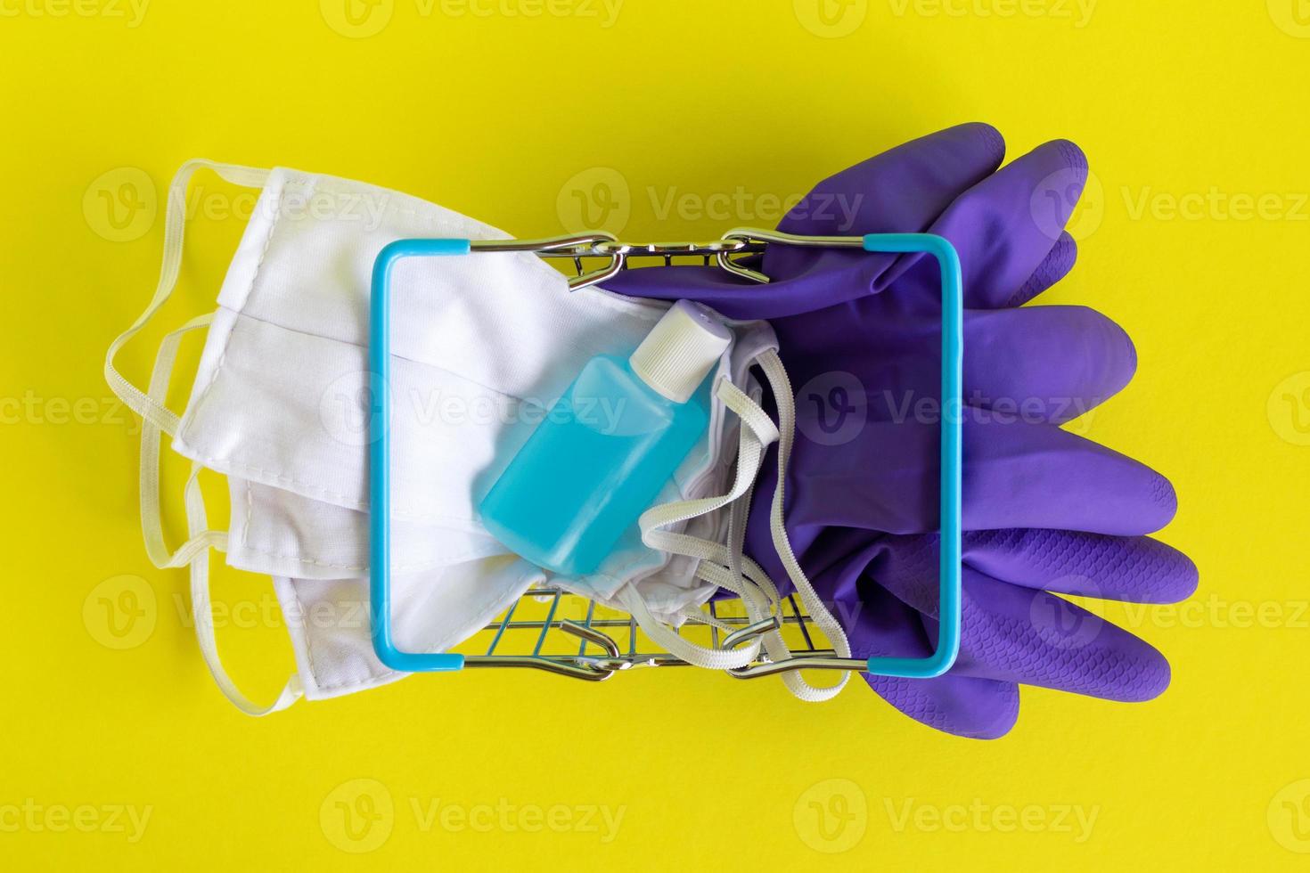 tumbado en la cesta de la compra con máscaras faciales, un par de guantes médicos desechables y una botella de gel desinfectante para manos sobre fondo amarillo. concepto de compras en el supermercado en un nuevo estilo de vida normal. foto