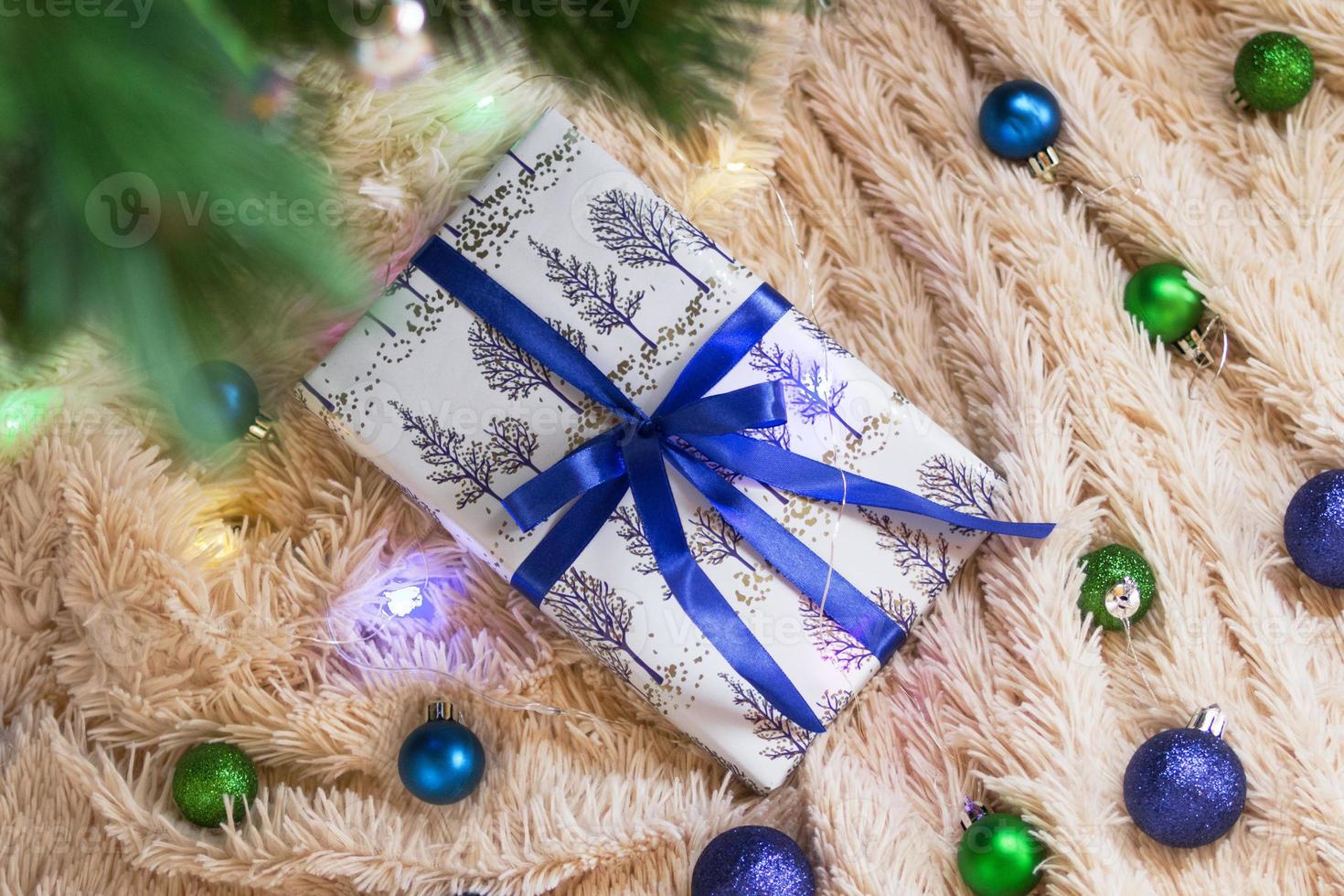Gift wrapped on white paper with blue ribbon on a background of beige plush plaid with Christmas lights and green and blue Christmas decorations, under branch of Christmas tree, top view. photo