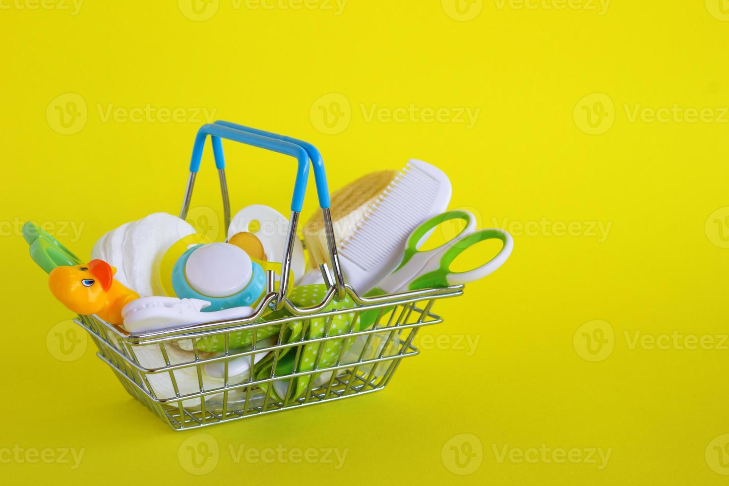 Shopping basket with baby care items - scissors, hairbrushes, pacifiers, thermometer, cotton pads, pacifier holders and nasal aspirator - on yellow background. photo