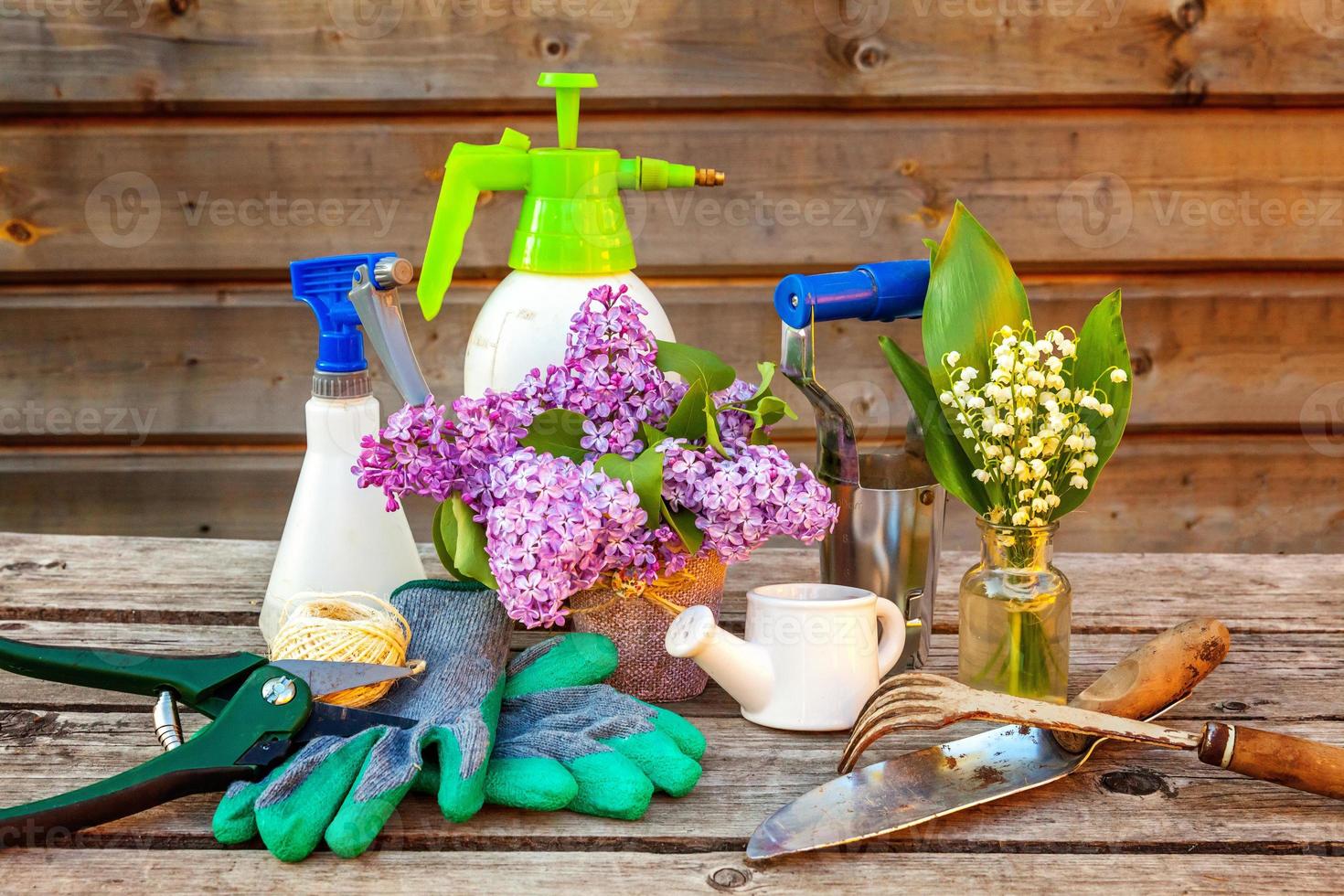 Gardening tools, watering can, shovel, spade, pruner, rake, glove, lilac, lily of the valley flowers on vintage wooden table. Spring or summer in garden, eco, nature, horticulture hobby concept. photo