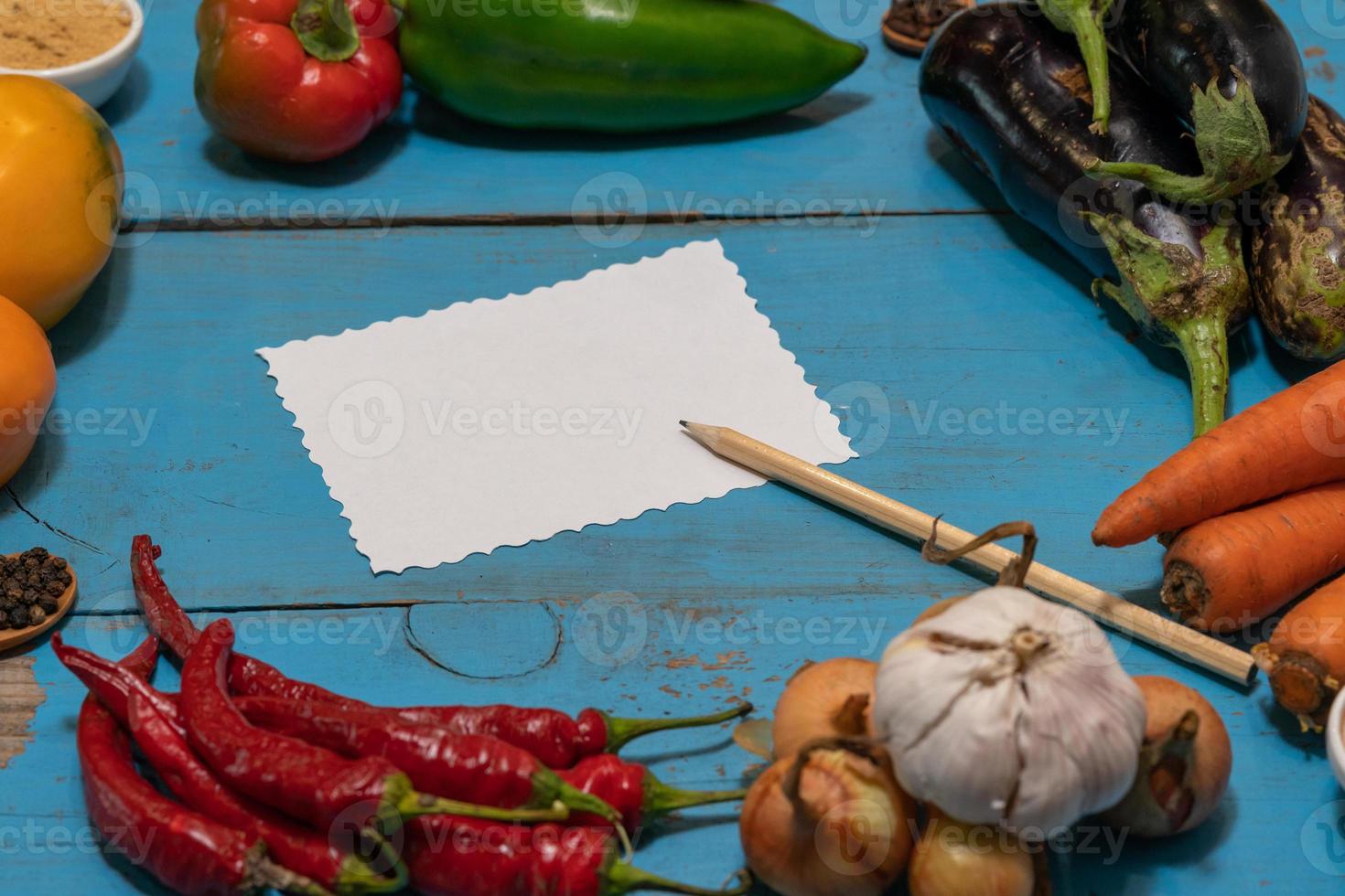 Vegetables are laid out around a sheet of paper and a pencil. Empty space for text. Vegetables, empty blank for recipe on a blue background. photo