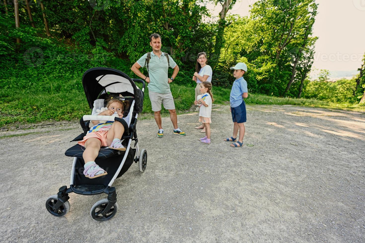 Family with three kids walking at park, baby girl hold toy in sitting stroller. photo