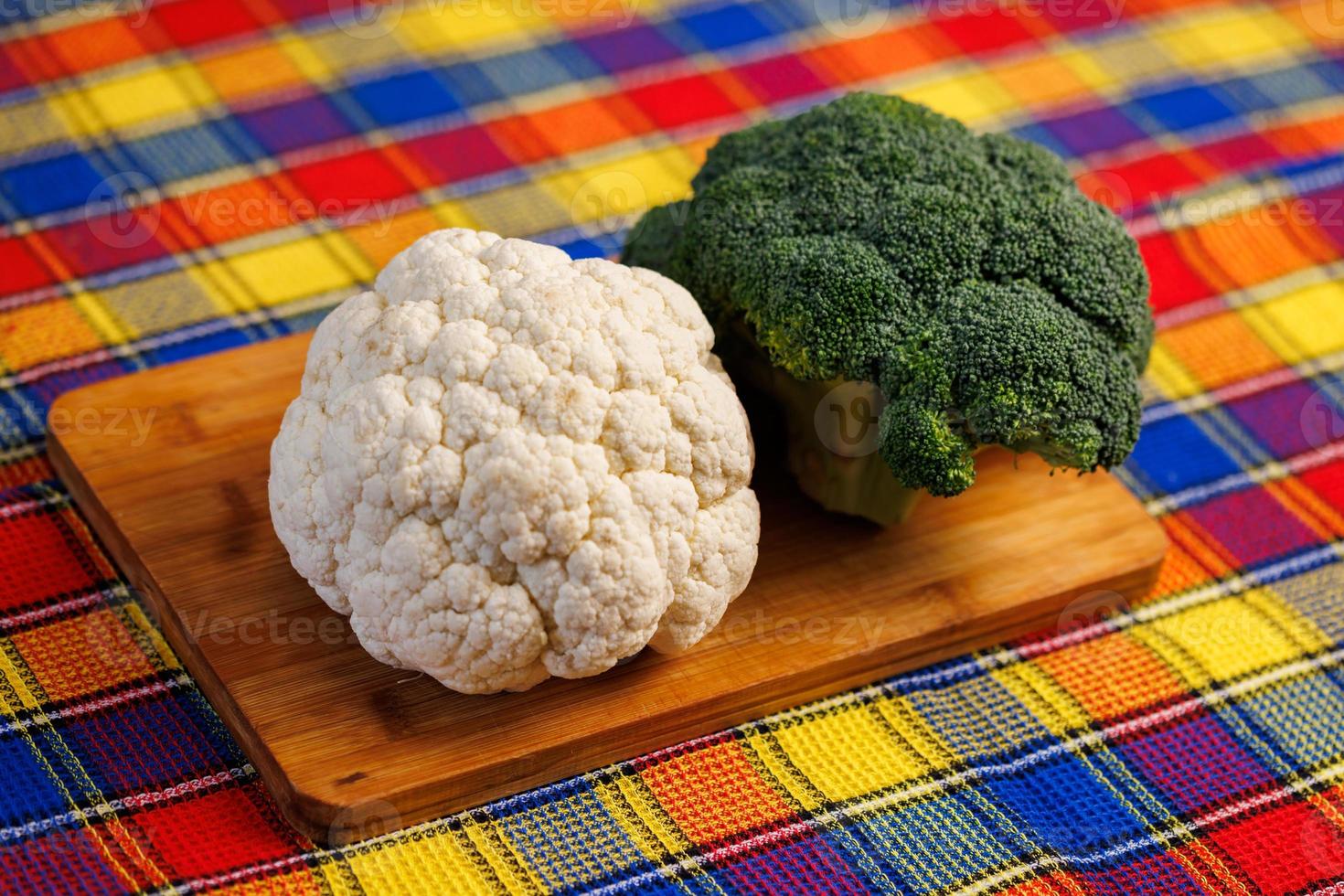 a head of broccoli and a head of cauliflower lie on a cutting board on the table with colorful towel underneath photo