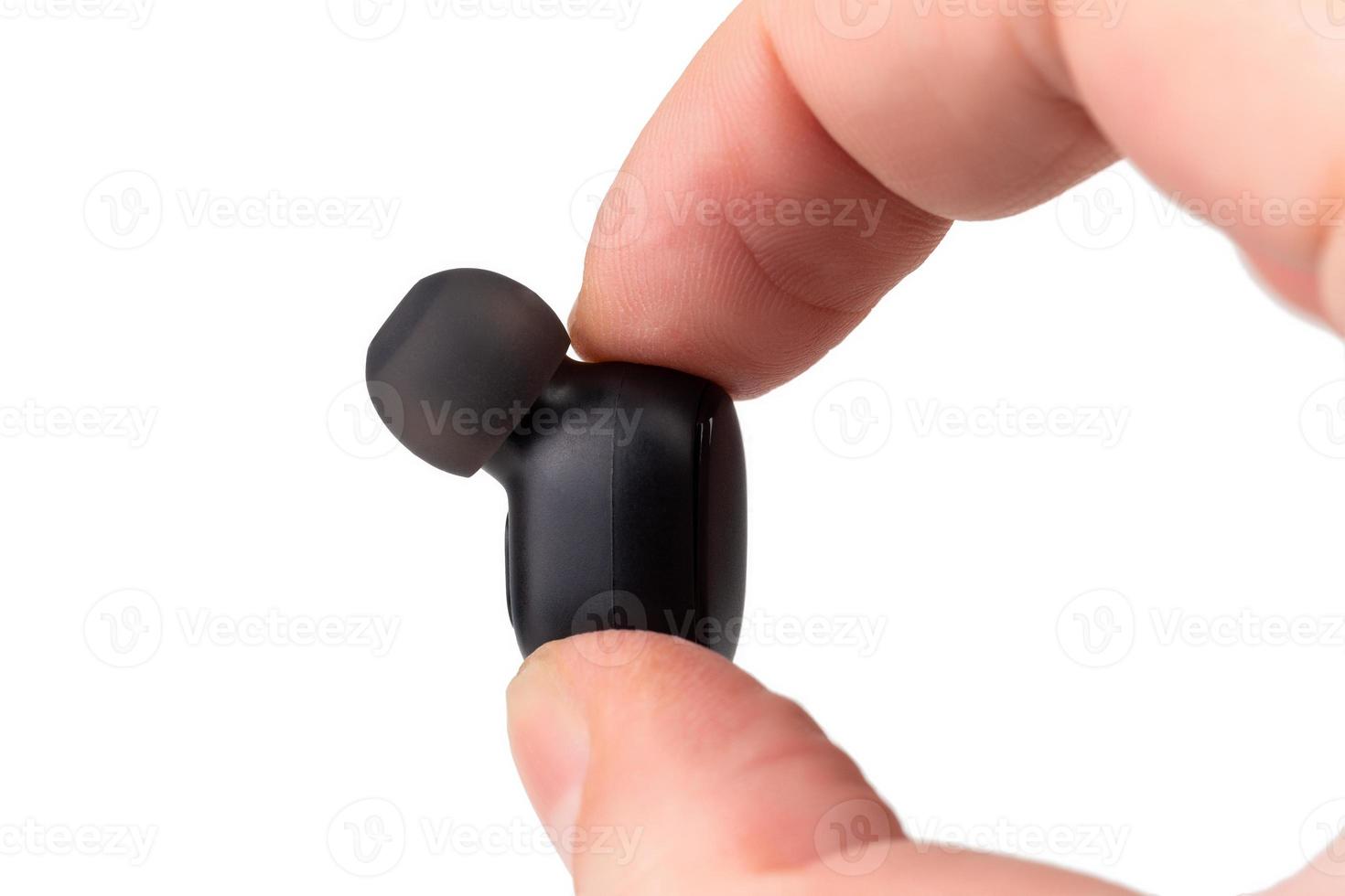 two caucasian fingers holding a black wireless earbud - close-up with selective focus on white background photo