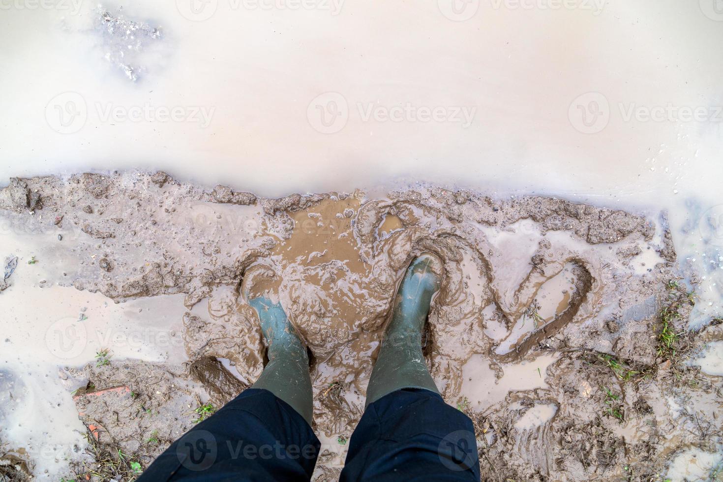 feet in green rubber boots stands in wet brown mud puddle directly above view photo