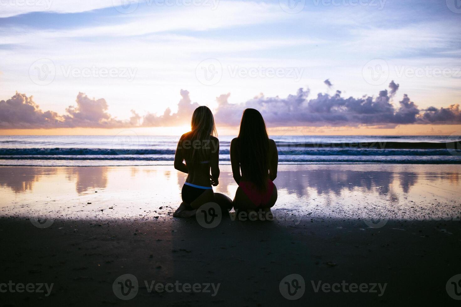 A dim shot of two women in bikini kneeling on a wet beach and admiring the sunset photo