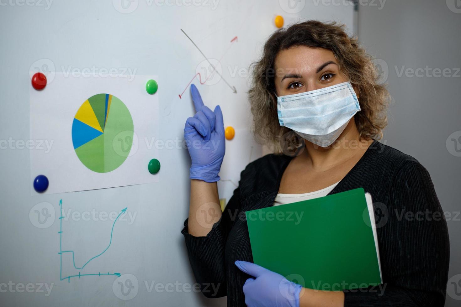 A young cheerful employee, a corporate lawyer, a woman in a mask and gloves working in an office shows paper accounting documents on a marker board. Protection in the office from viruses. photo