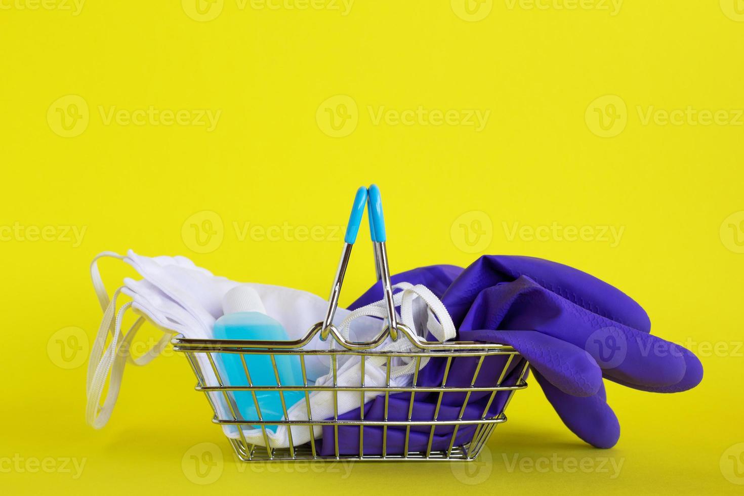 máscaras faciales, un par de guantes médicos desechables y una botella de gel desinfectante para manos en una pequeña cesta de compras sobre fondo amarillo con espacio para copiar. concepto de compras en el supermercado en un nuevo estilo de vida normal. foto