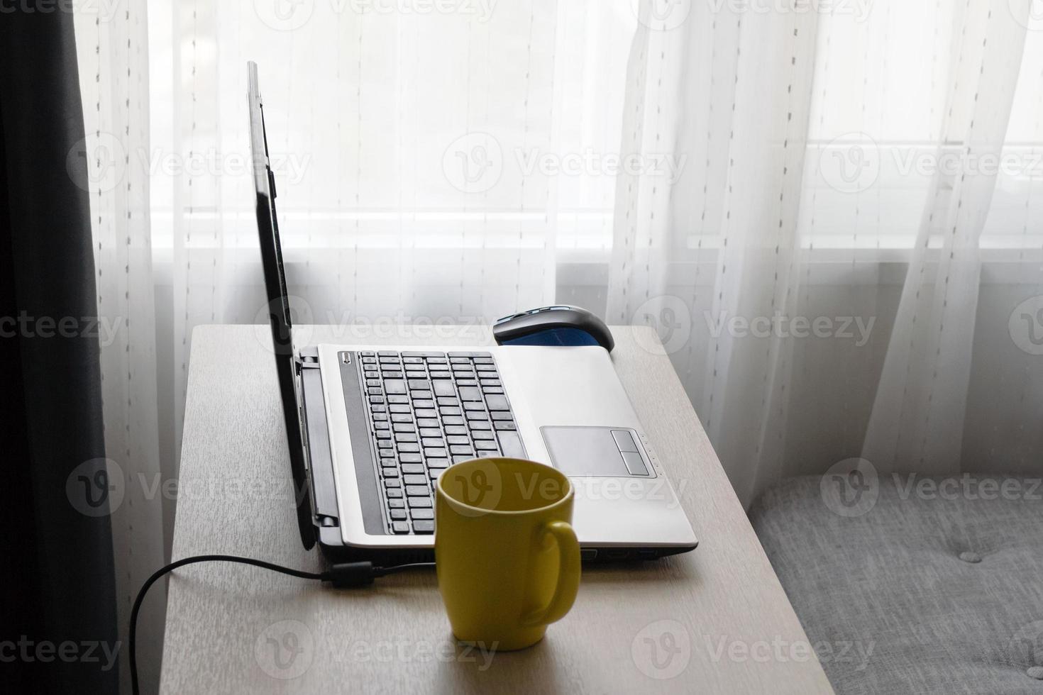 concepto de oficina en casa. trabajar desde casa en la sala de estar. portátil y taza de bebida caliente en la mesa de madera cerca del sofá y la ventana. foto