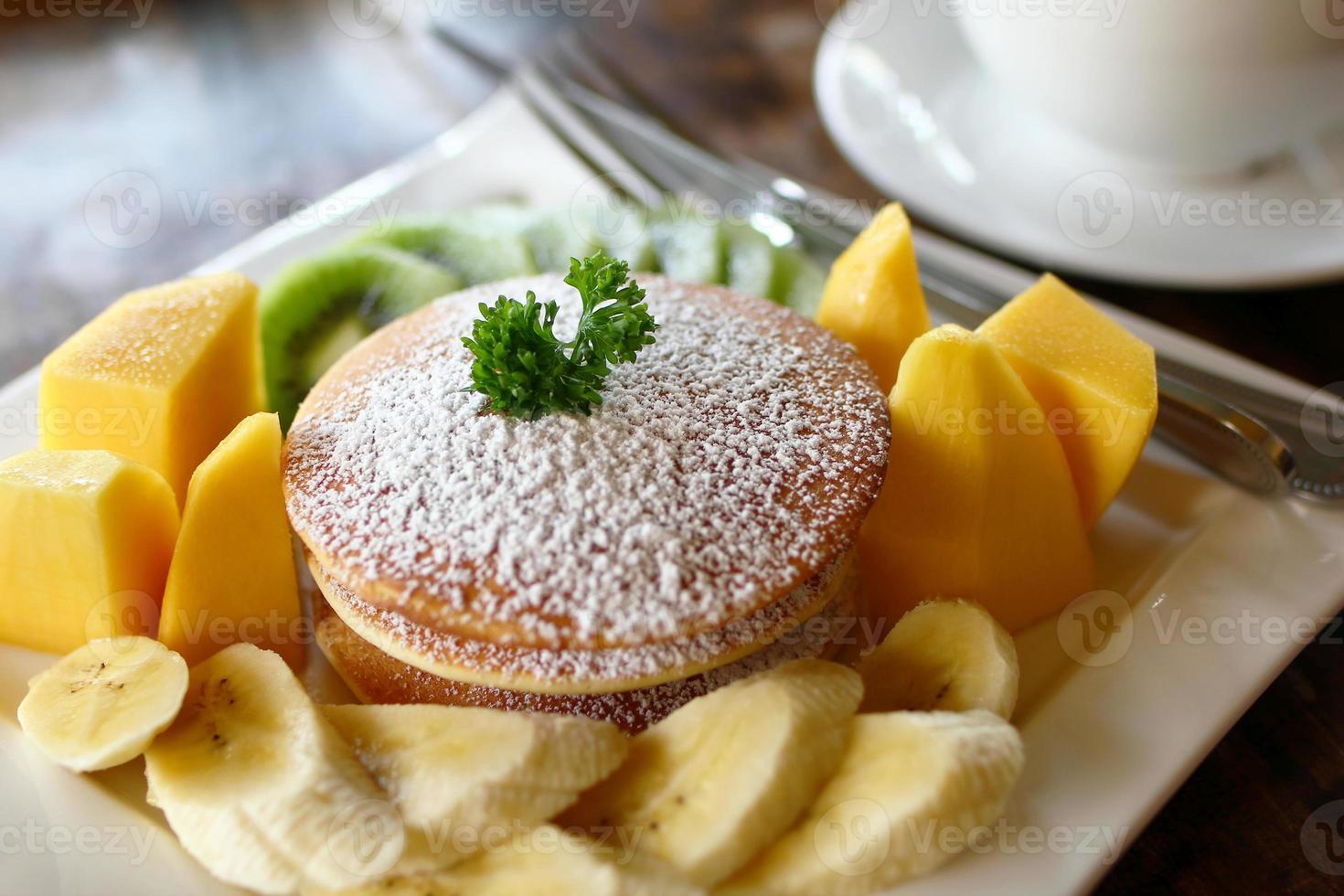 Plate with pancakes with fresh fruits - mango, banana and kiwi - decorated powdered sugar and leaf of mint on a table in cafe with cup of coffee on a background. photo