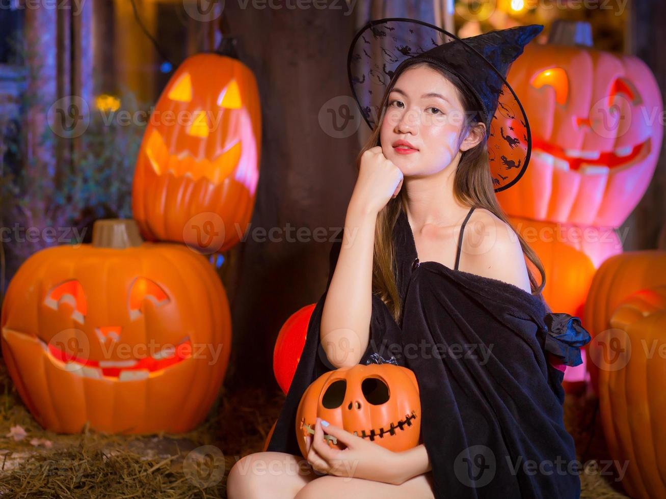 la mujer asiática usa un abrigo negro fantasma y se maquilla como un fantasma de bruja mientras mira a la cámara y sostiene una linterna de cabeza de calabaza en sus manos por la noche en un escenario al aire libre contra un fondo. foto
