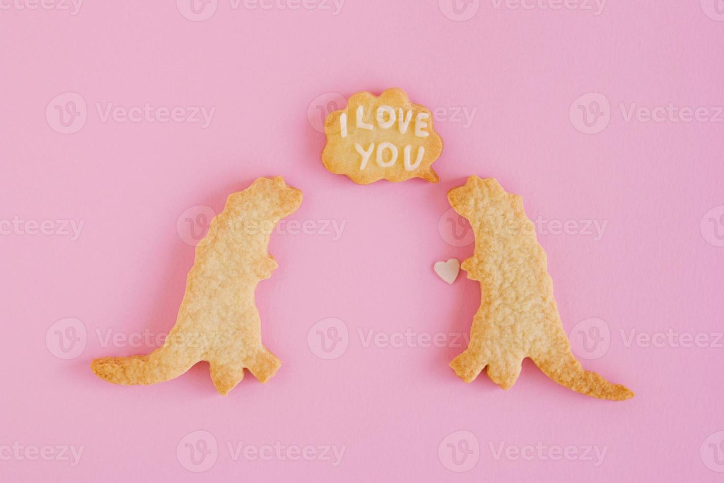 galletas de mantequilla caseras con glaseado blanco sobre fondo rosa, vista superior. dos dinosaurios con nube de llamada con texto - te amo foto