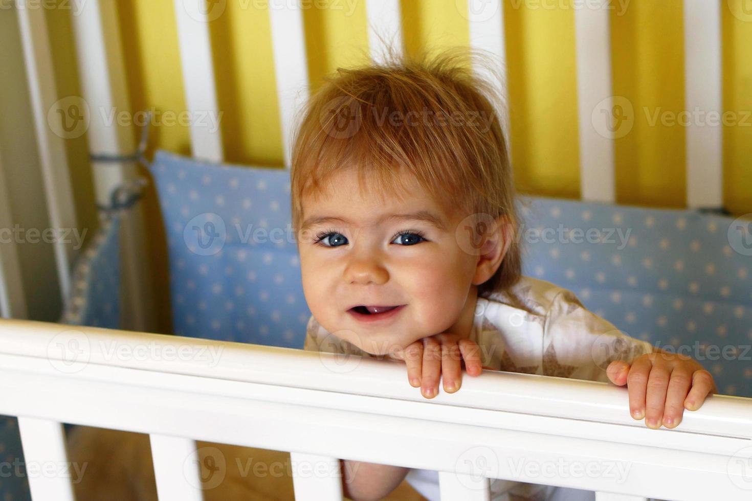 Happy one year old girl with blue eyes and blond after waking up in the morning in her bed. photo