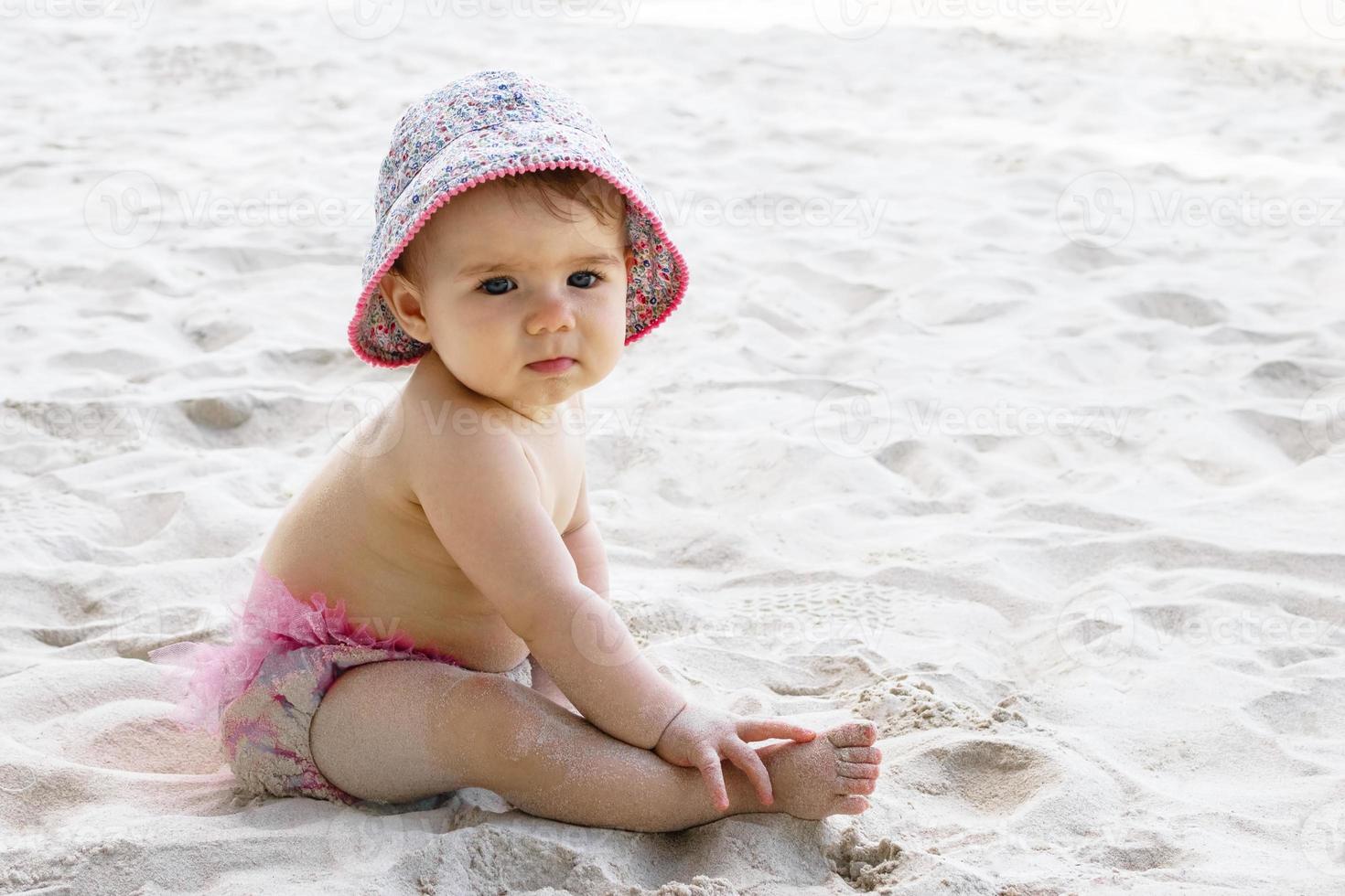 linda niñita con bañador rosa y panamá sentada en la arena de la playa. foto