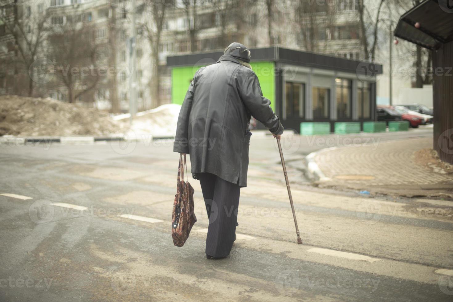 Old woman walks down road in black jacket. Pensioner in Russia on street. photo
