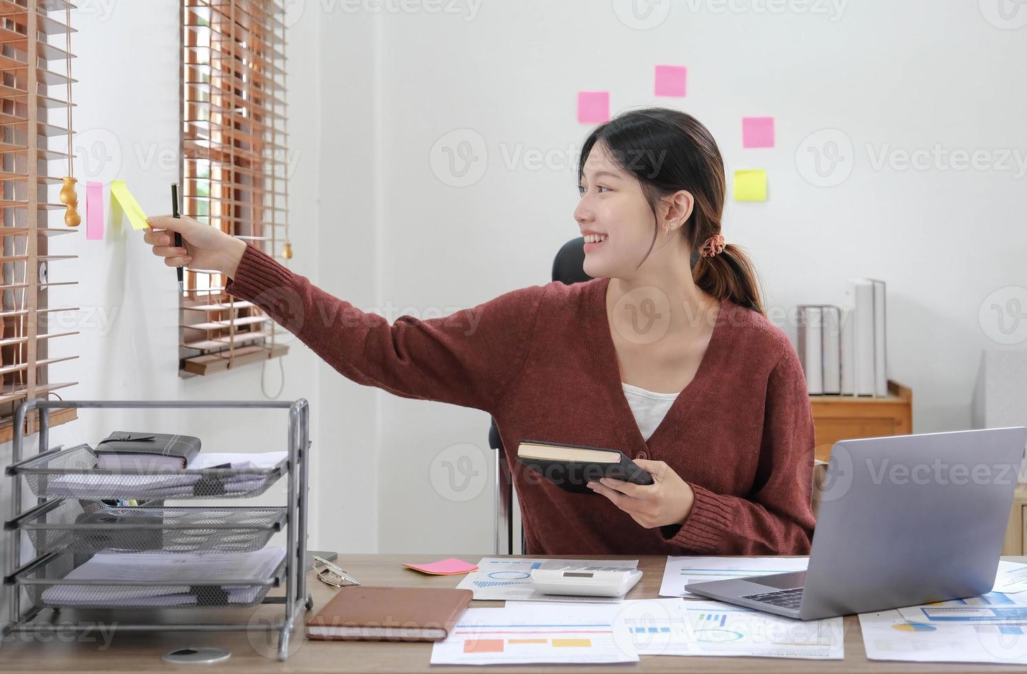 mujer asiática de negocios tomando nota en una nota adhesiva, chica atractiva que trabaja con una computadora portátil foto