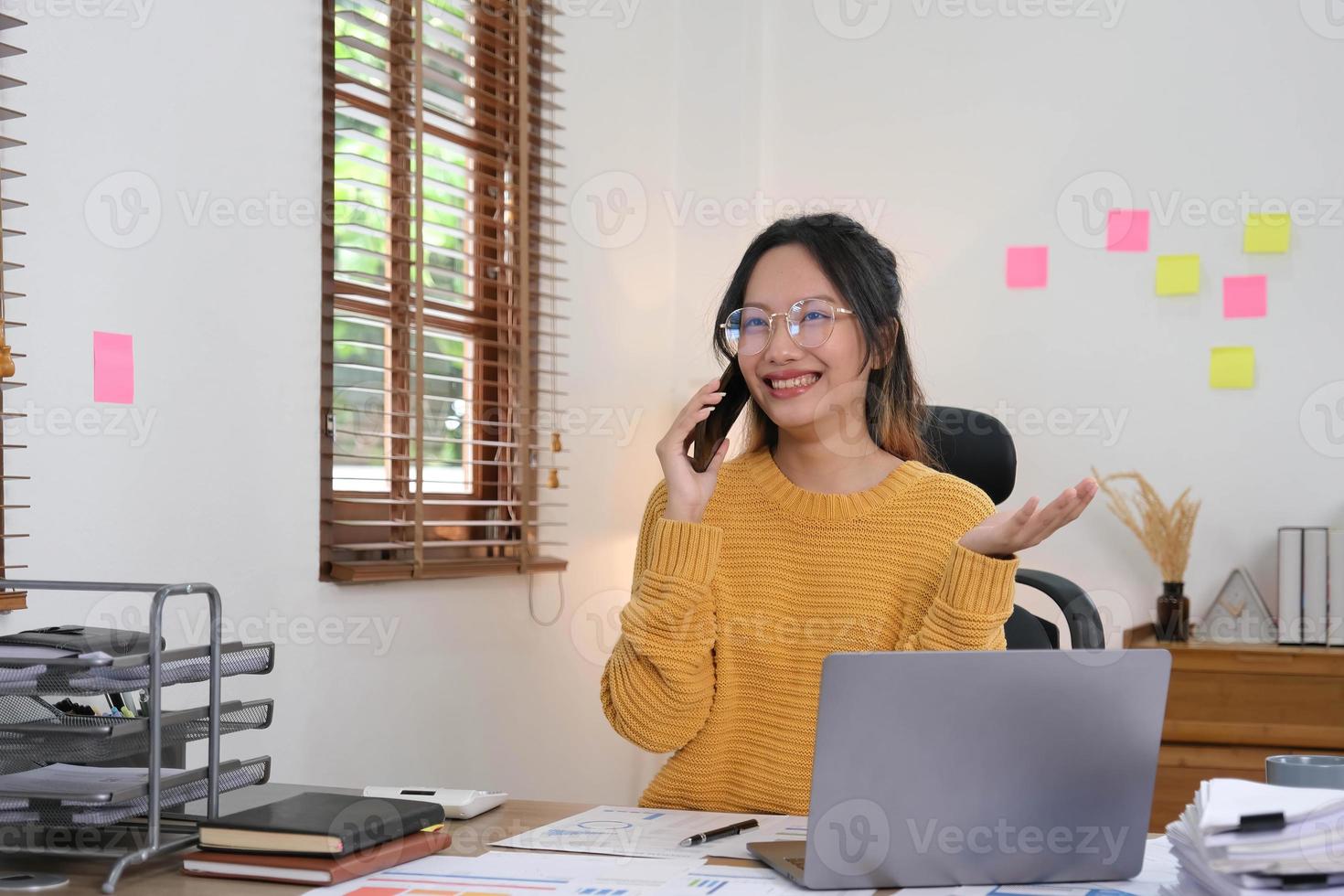 atractiva y sonriente mujer de negocios asiática está hablando por un teléfono móvil con un cliente con una computadora portátil en la oficina. foto