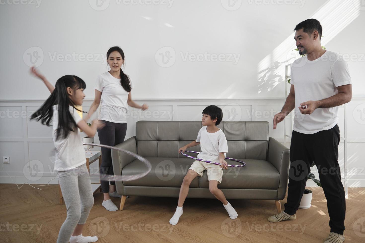 Happy wellness Asian Thai family, parents, and children are fun playing hula hoops together, fitness training and healthy exercise in white living room, domestic home lifestyle, and weekend activity. photo