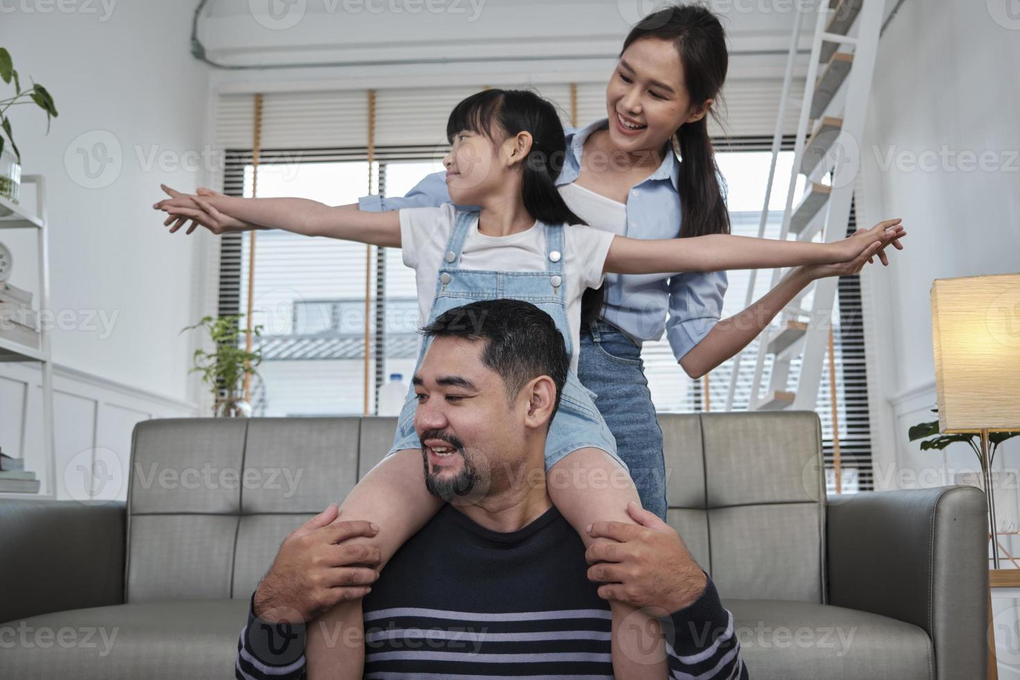 familia asiática tailandesa junta, papá juega y bromea con su hija y su madre cargando y sosteniendo a la niña sobre los hombros en la sala de estar de la casa, tiempos de ocio felices, fin de semana encantador, estilo de vida doméstico de bienestar. foto