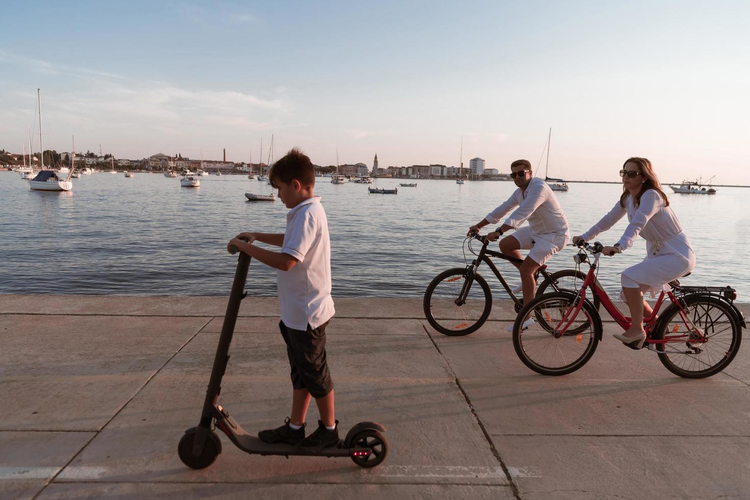 familia feliz disfrutando juntos de una hermosa mañana junto al mar, padres montando en bicicleta y su hijo montando una scooter eléctrica. enfoque selectivo foto