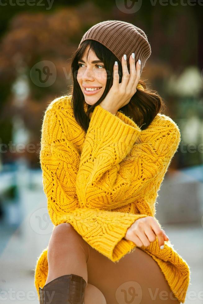 Beautiful European brunette dressed in a yellow woolen sweater and hat outside. The beautiful girl who wears thick stylish autumn clothes in cool weather. photo