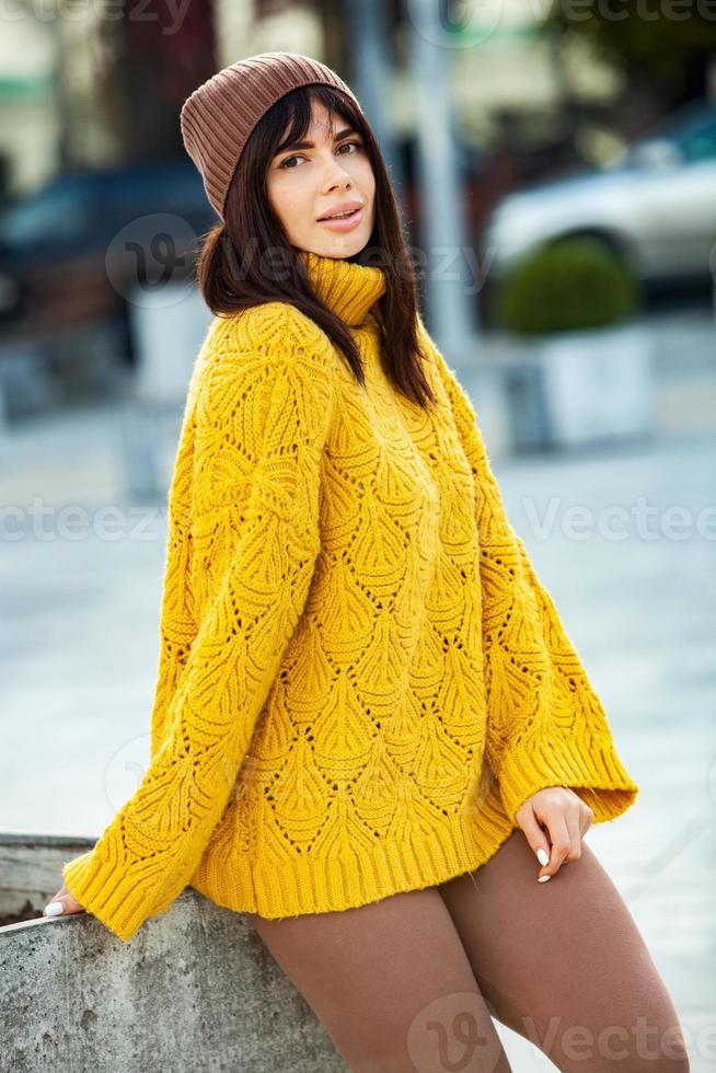 Beautiful European brunette dressed in a yellow woolen sweater and hat outside. The beautiful girl who wears thick stylish autumn clothes in cool weather. photo