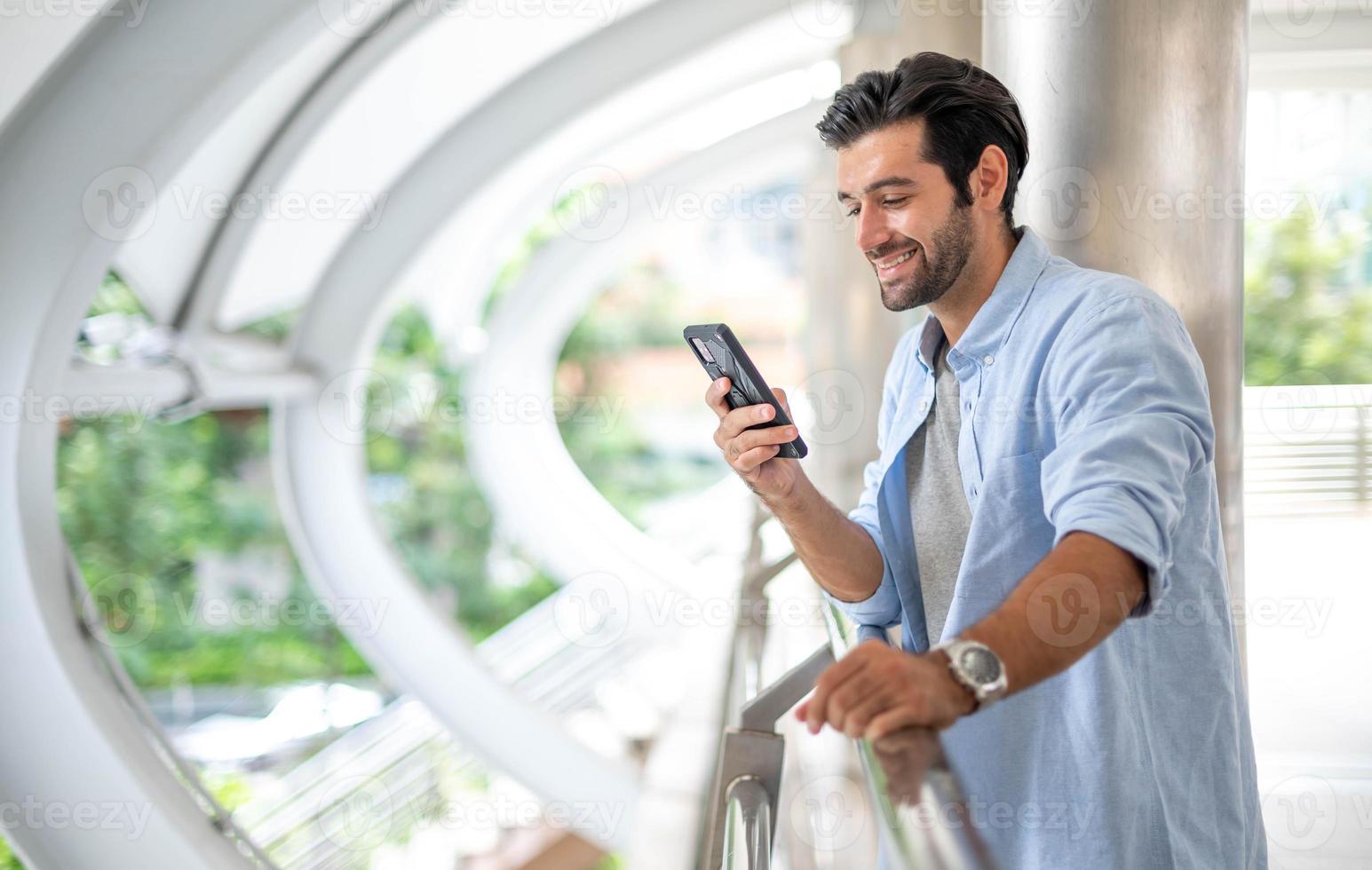 joven caucásico usando un teléfono inteligente y sonriendo mientras espera a su amigo afuera. el hombre que sostiene y usa el teléfono celular para buscar datos y redes sociales en Internet. foto