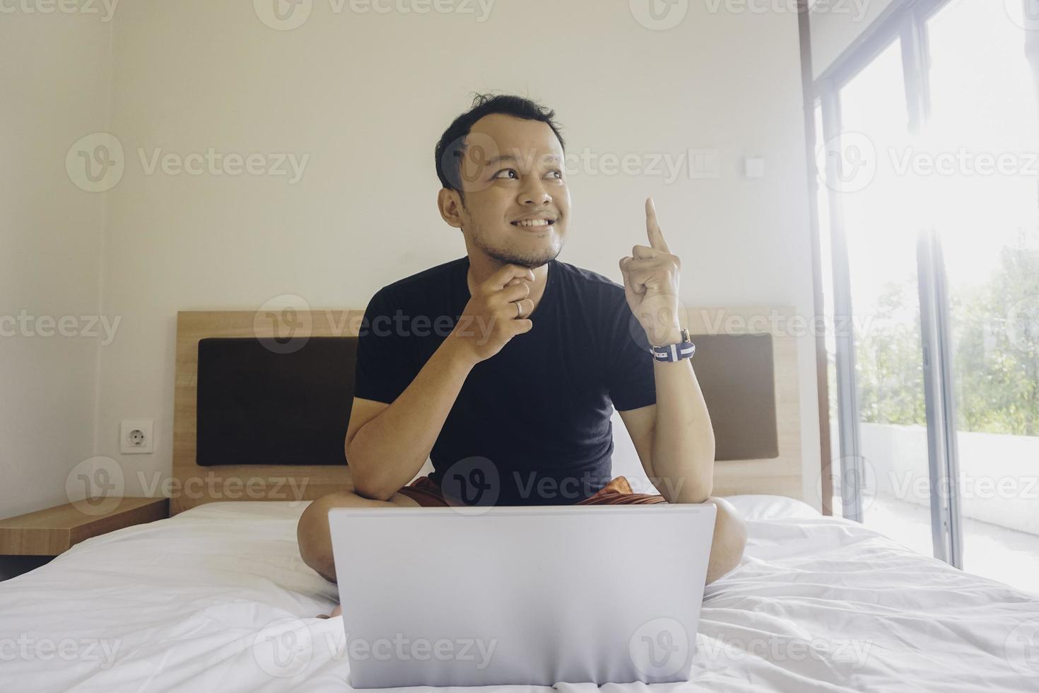 Thoughtful young Asian man showing idea gesture by pointing upwards works remotely behind a laptop photo