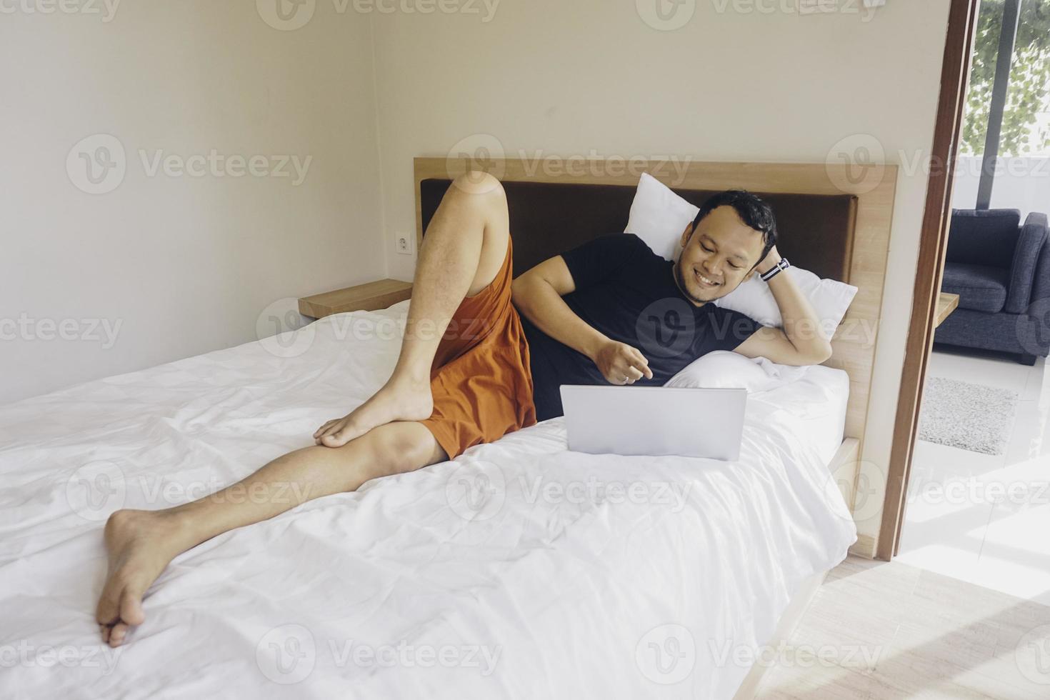 Smiling young man working from the bed. Lying while checking at the laptop. photo