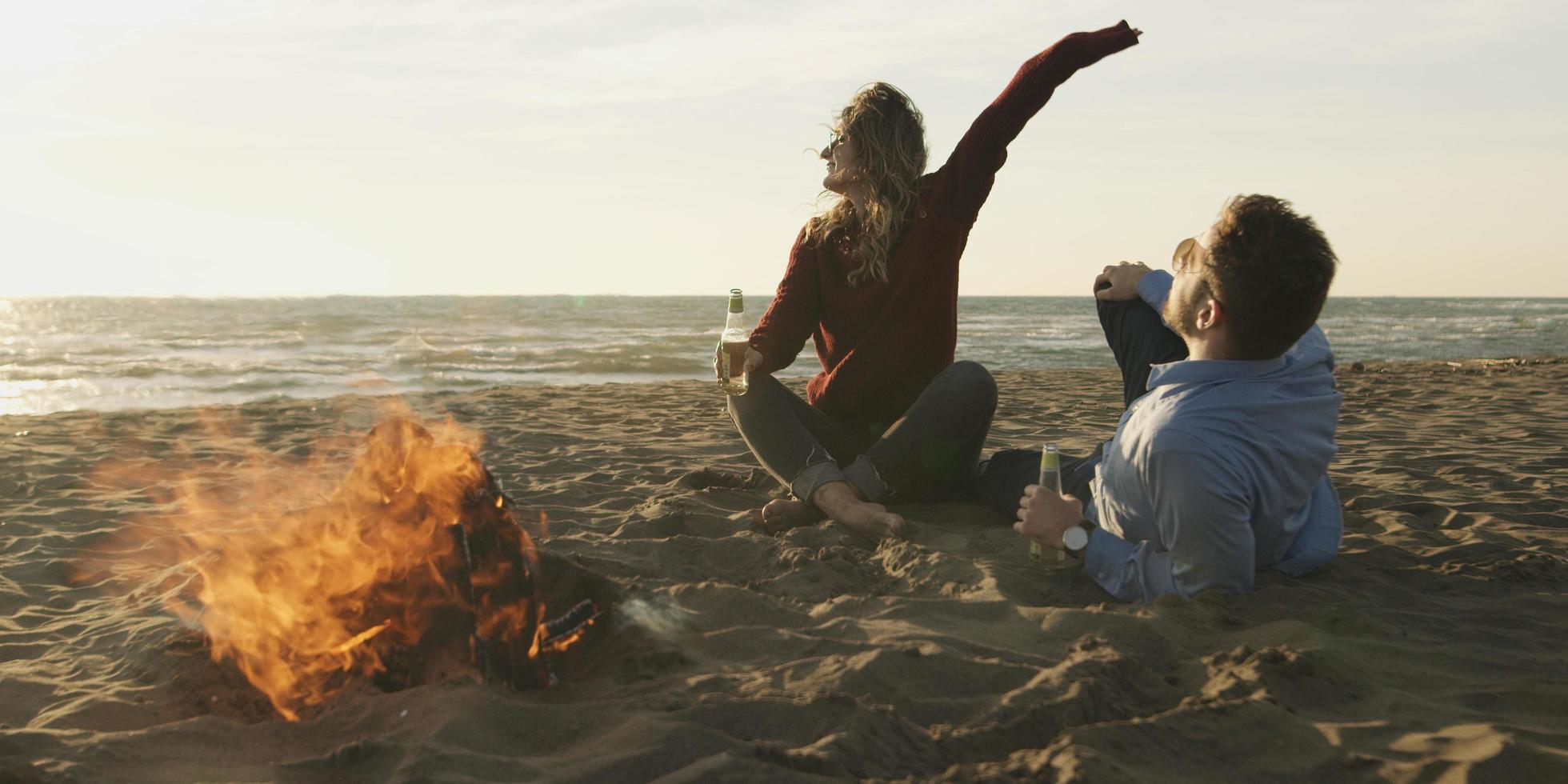 amorosa pareja joven sentada en la playa junto a la fogata bebiendo cerveza foto