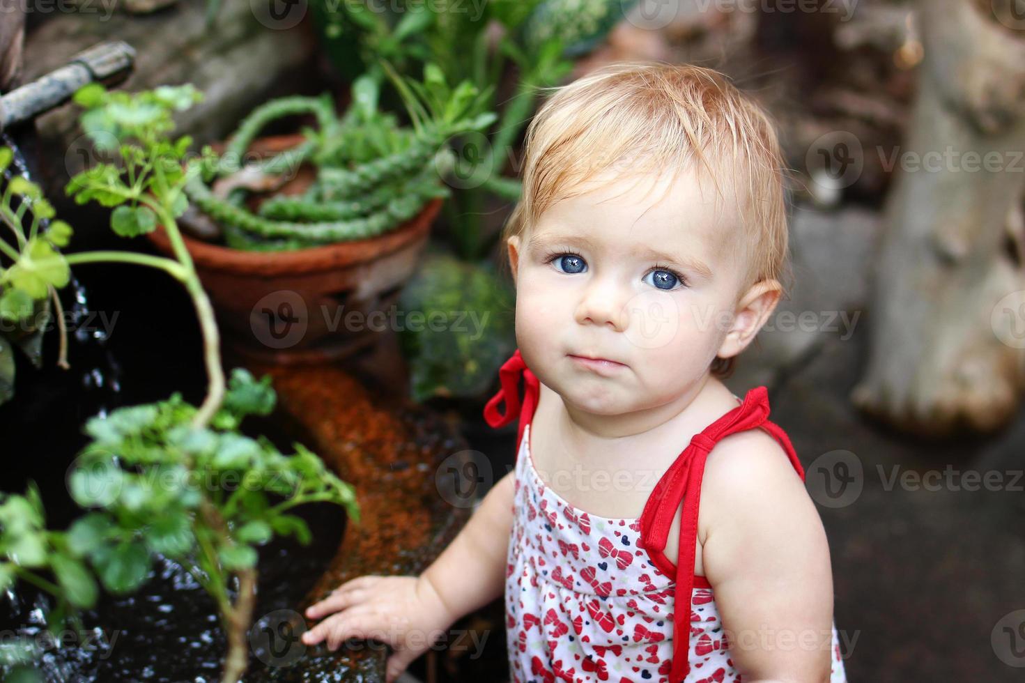 Portrait of little girl with blond hair and blue eyes in sundress in garden with pot plants and waterfall. photo