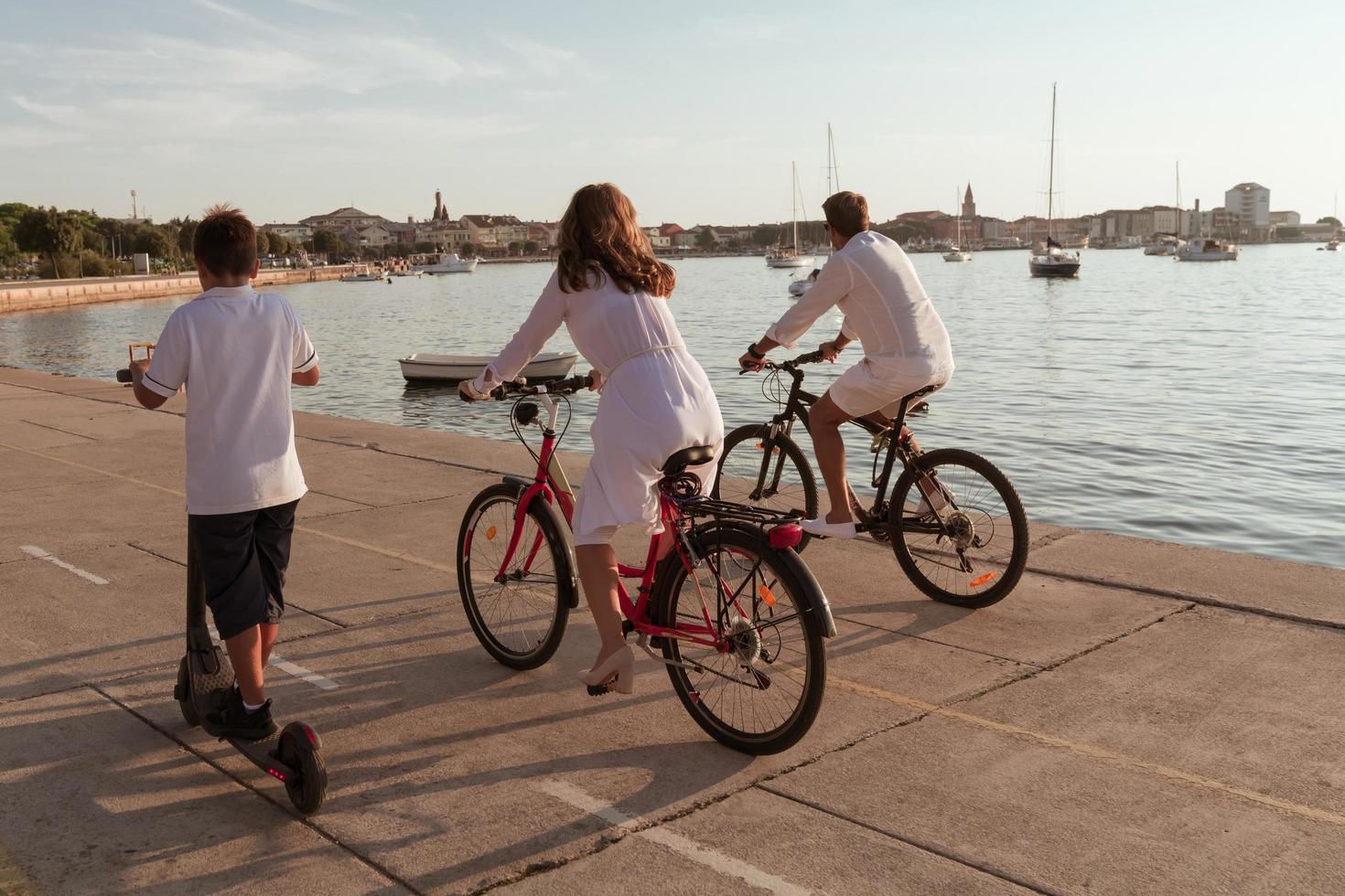 familia feliz disfrutando juntos de una hermosa mañana junto al mar, padres montando en bicicleta y su hijo montando una scooter eléctrica. enfoque selectivo foto