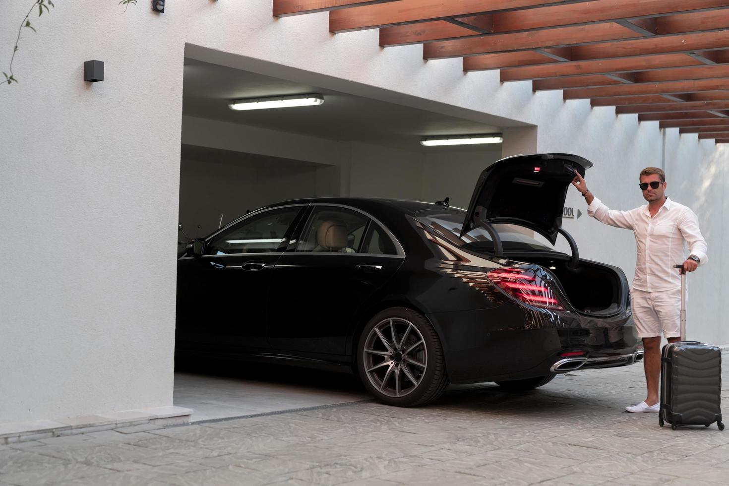 A senior man in a garage with glasses and modern casual clothes takes out his suitcases from the car. Selective focus photo